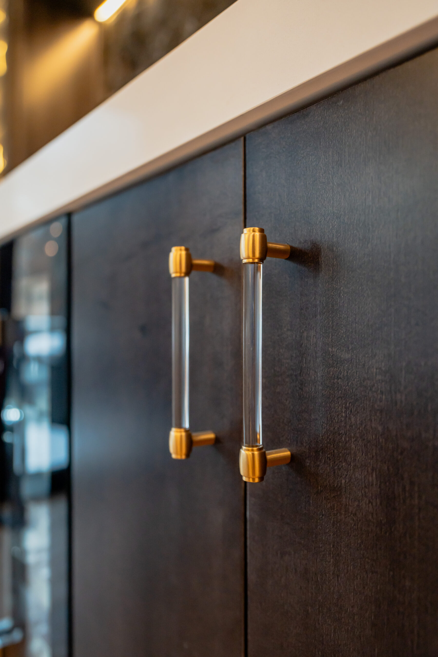 A close-up of modern clear acrylic cabinet handles with brushed gold accents on dark charcoal bar cabinetry.