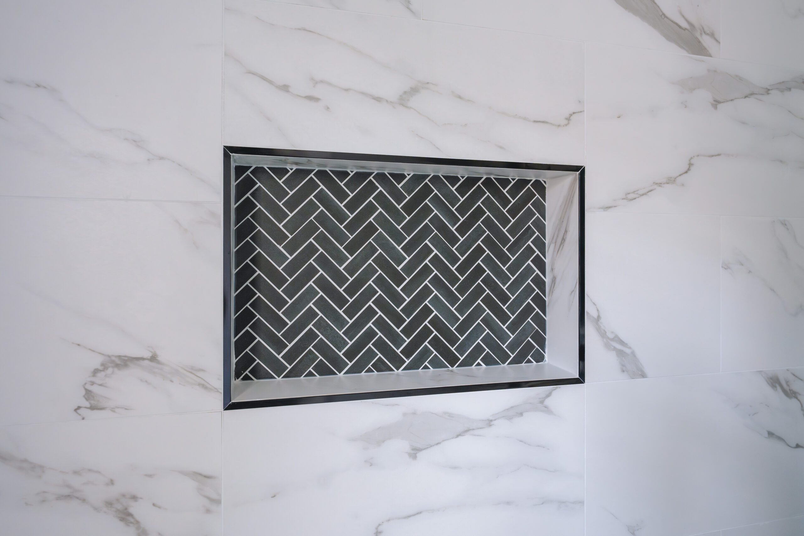 A recessed shower wall niche featuring a black herringbone tile pattern framed by large white marble-look tiles.