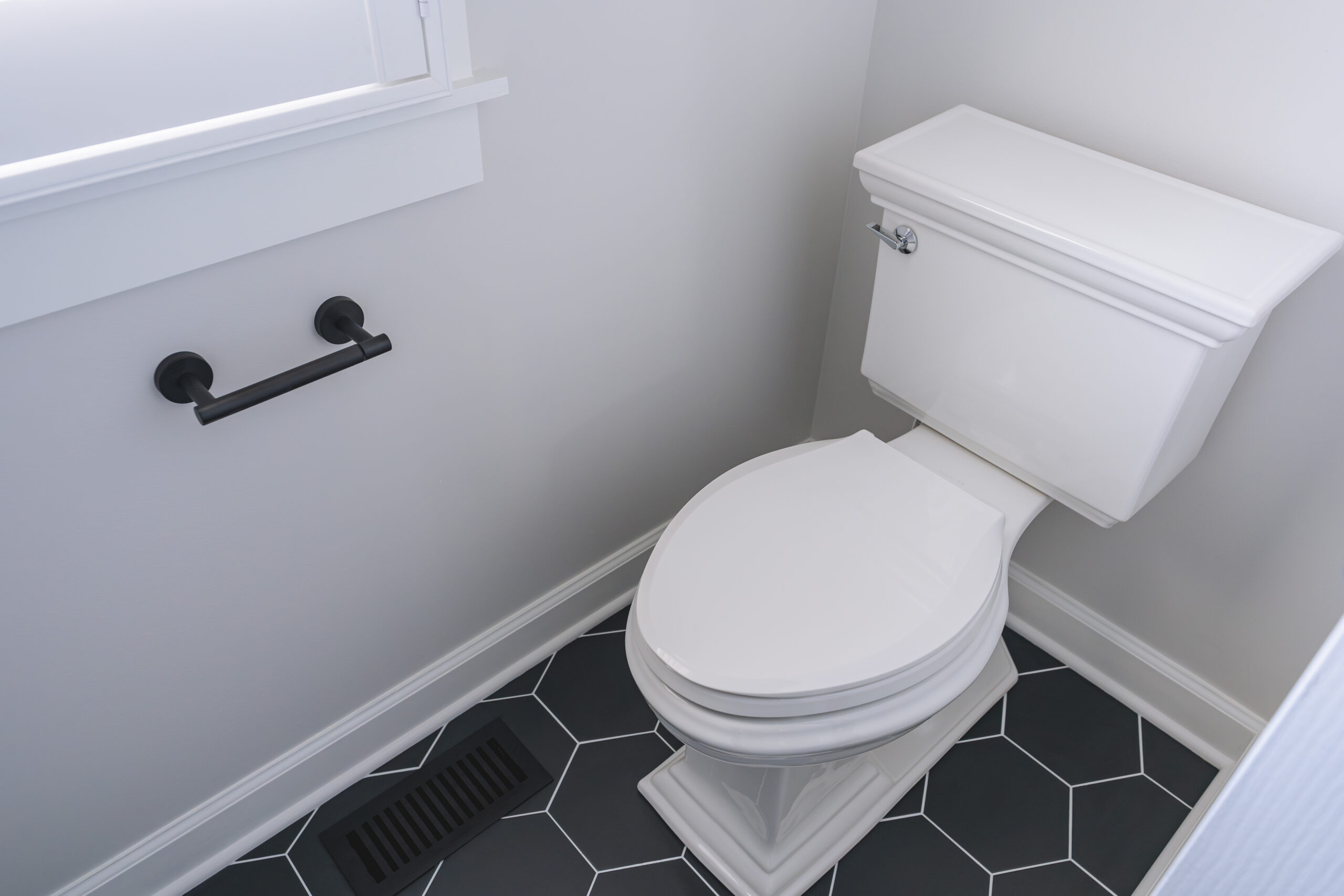A white traditional-style toilet set on black hexagon floor tiles with a black toilet paper holder.