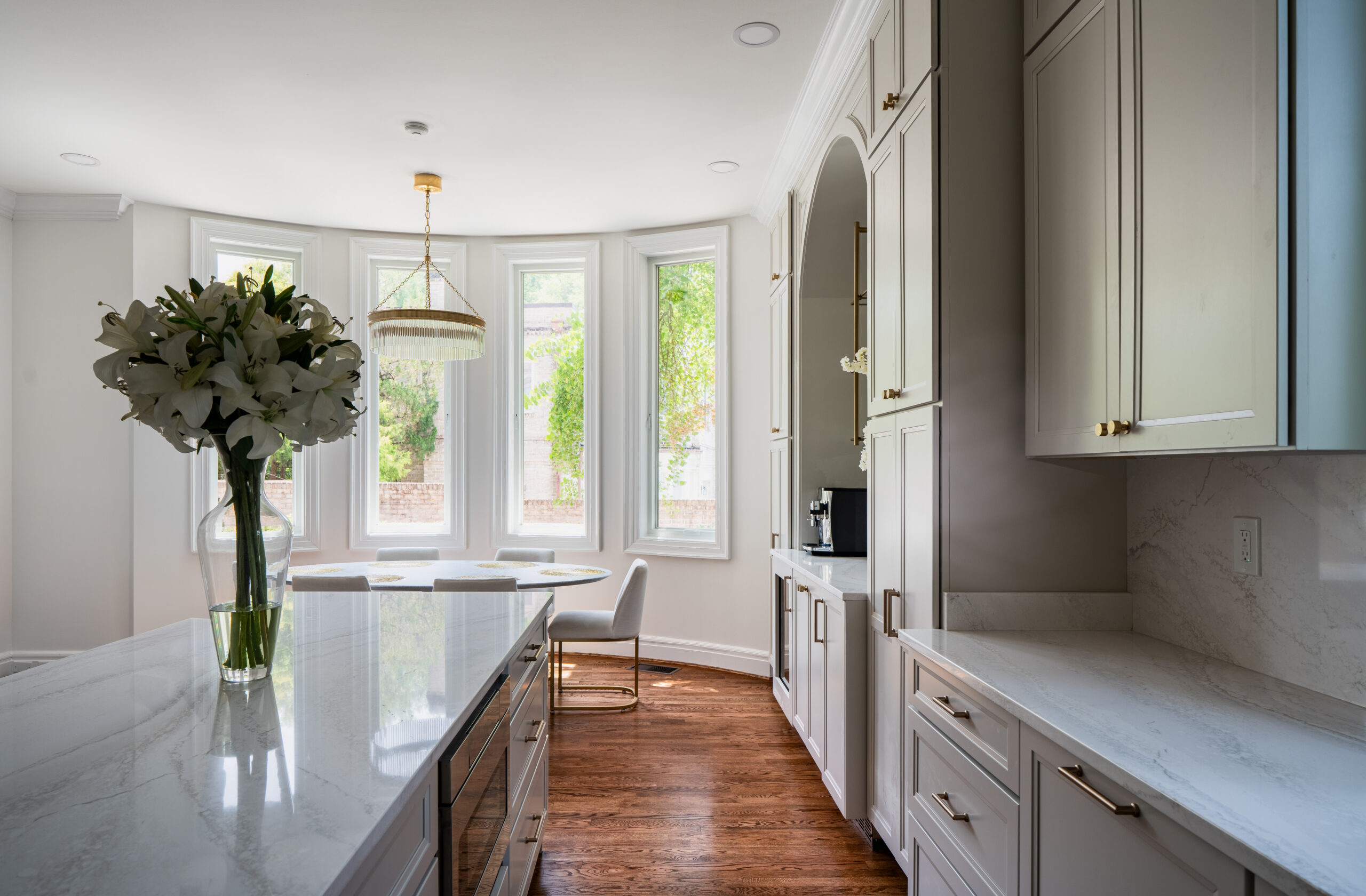 A sunlit dining area with an oval marble table and a crystal ring chandelier surrounded by large windows.