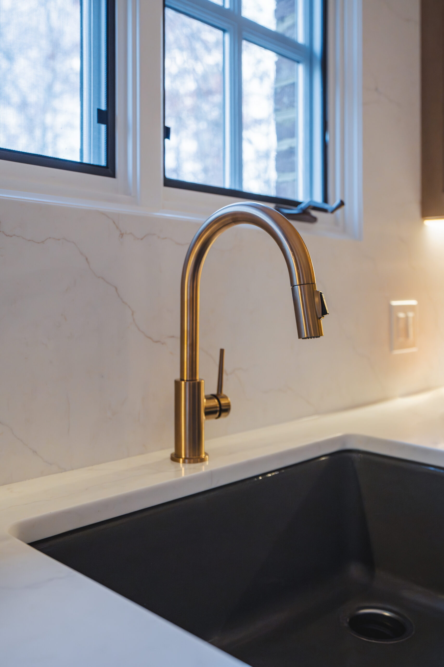 Close-up of a brushed gold gooseneck pull-down faucet over a matte black undermount farmhouse sink with a quartz backsplash.