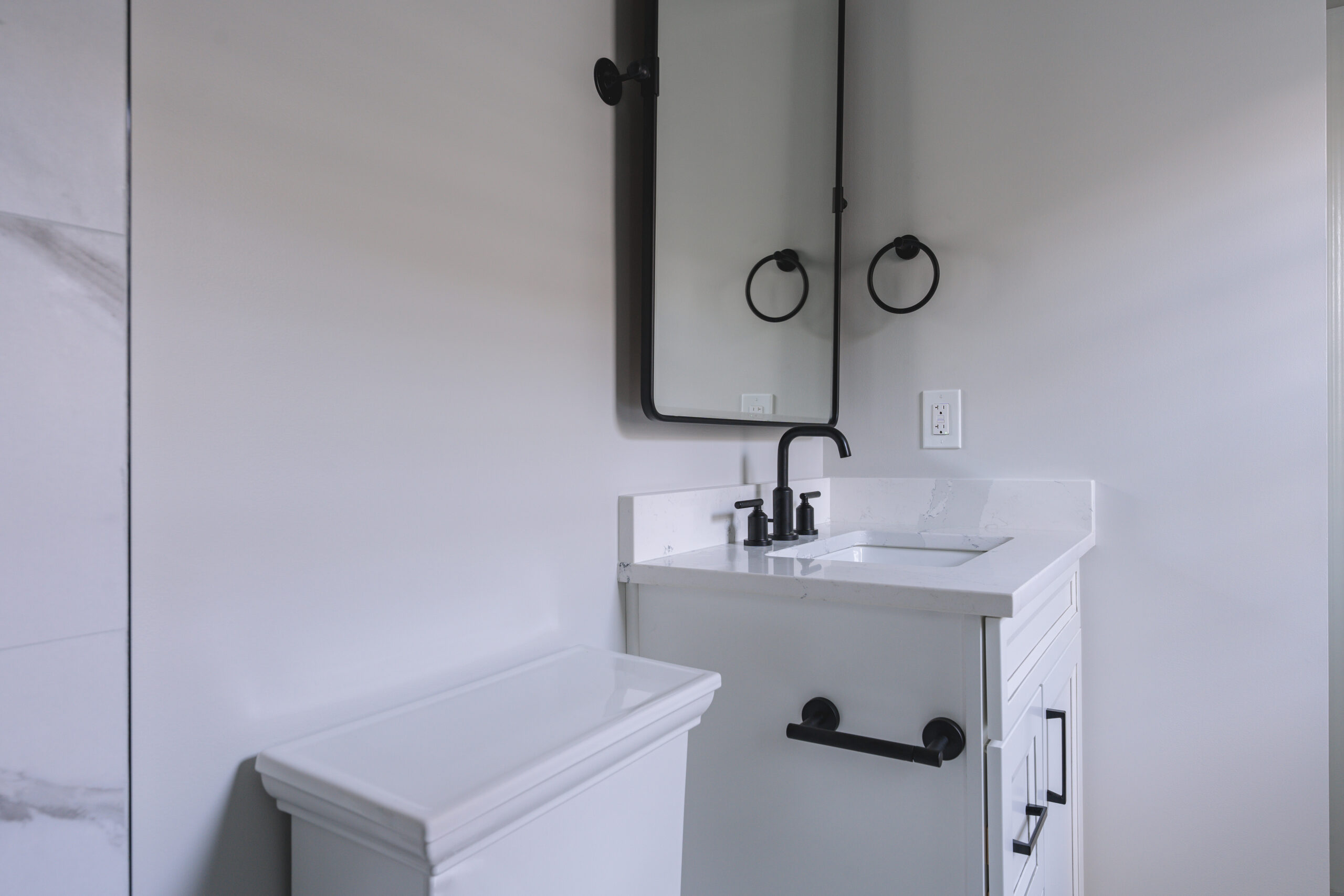 A compact white bathroom vanity with a black faucet, black-framed mirror, and matching towel ring.