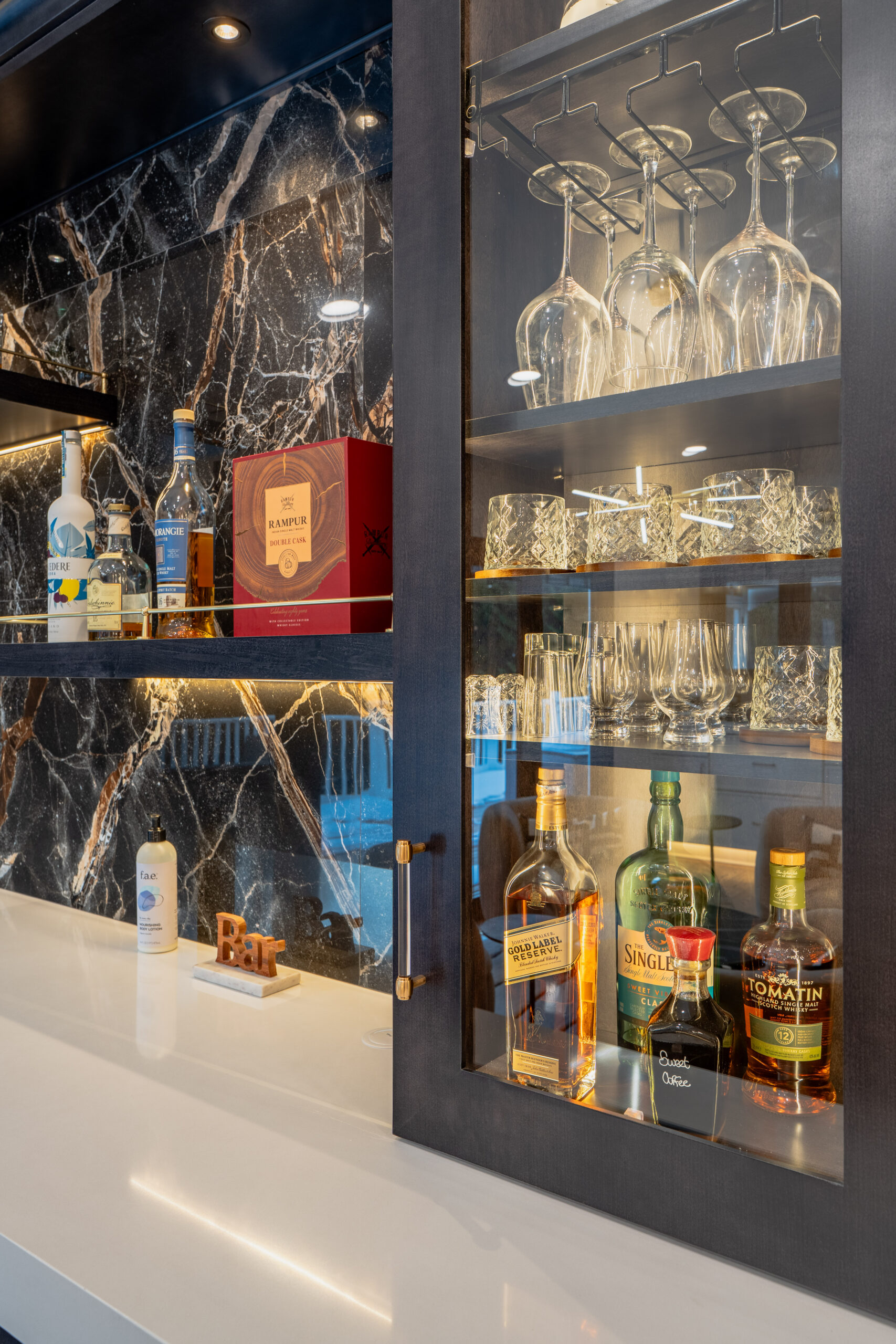A close-up of a dark wood home bar cabinet with glass shelves, hanging wine glasses, and a selection of premium liquor.
