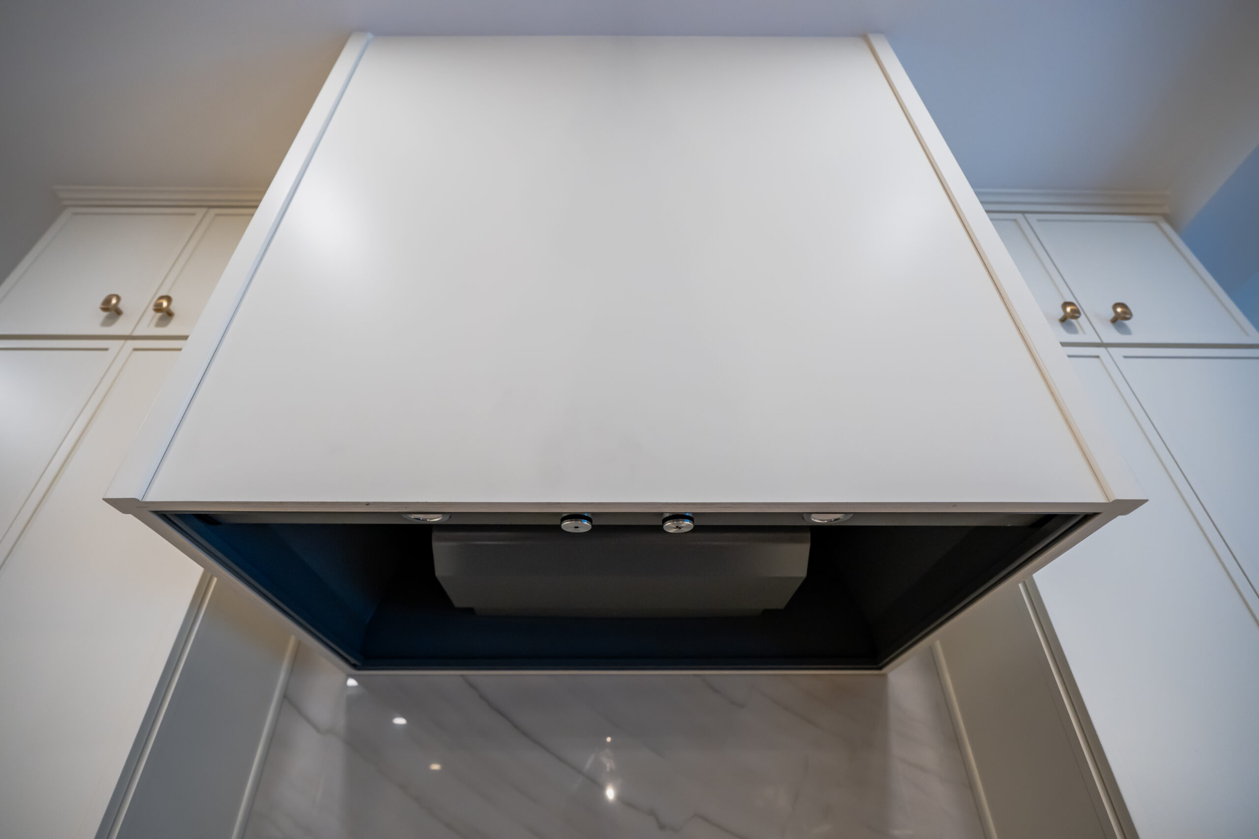 A low-angle view looking up into a large custom white range hood with professional-grade lighting and ventilation over a marble backsplash.