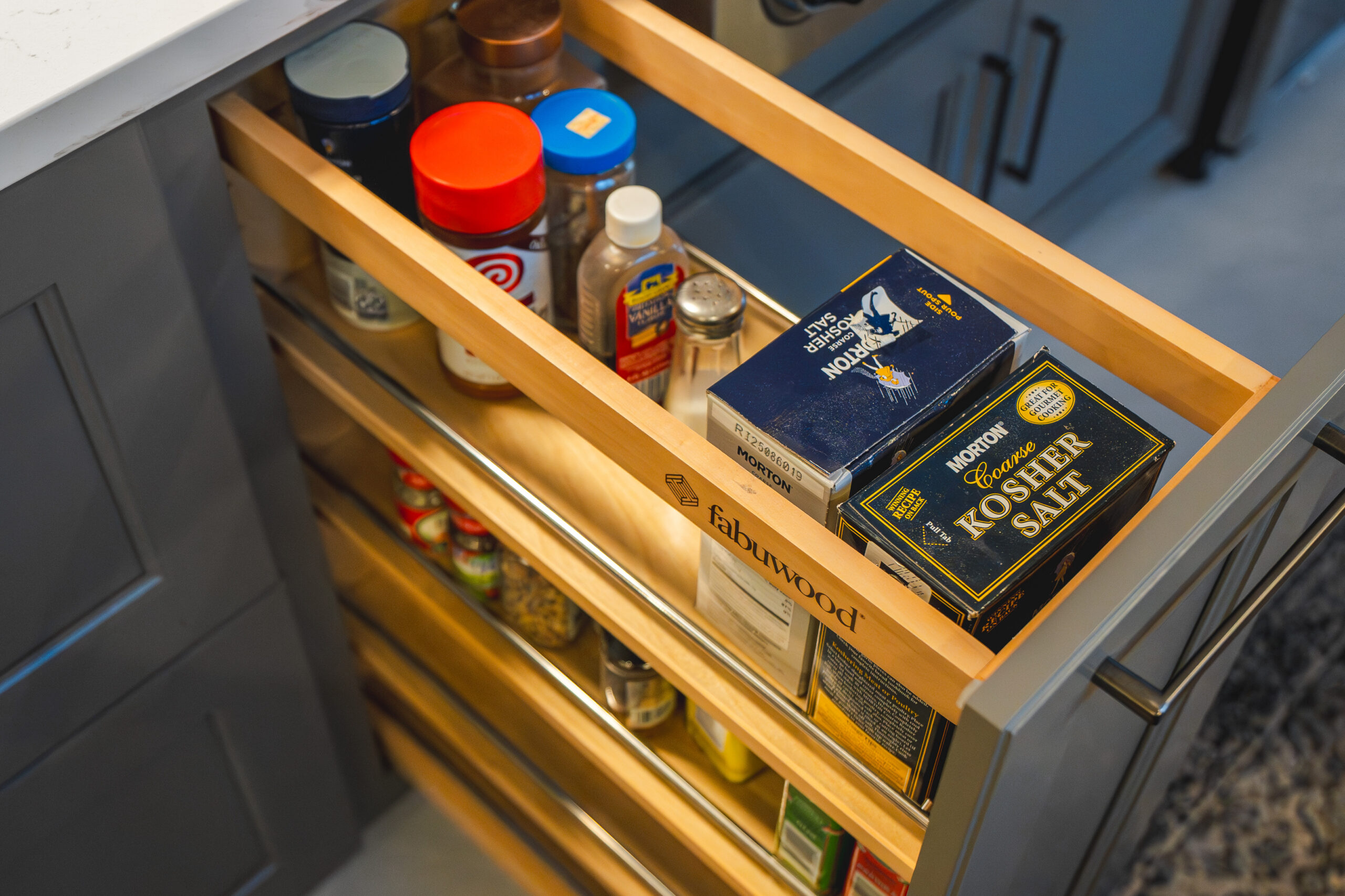 A wooden Fabuwood pull-out spice rack drawer showing organized spice containers and cooking salts inside a kitchen island.