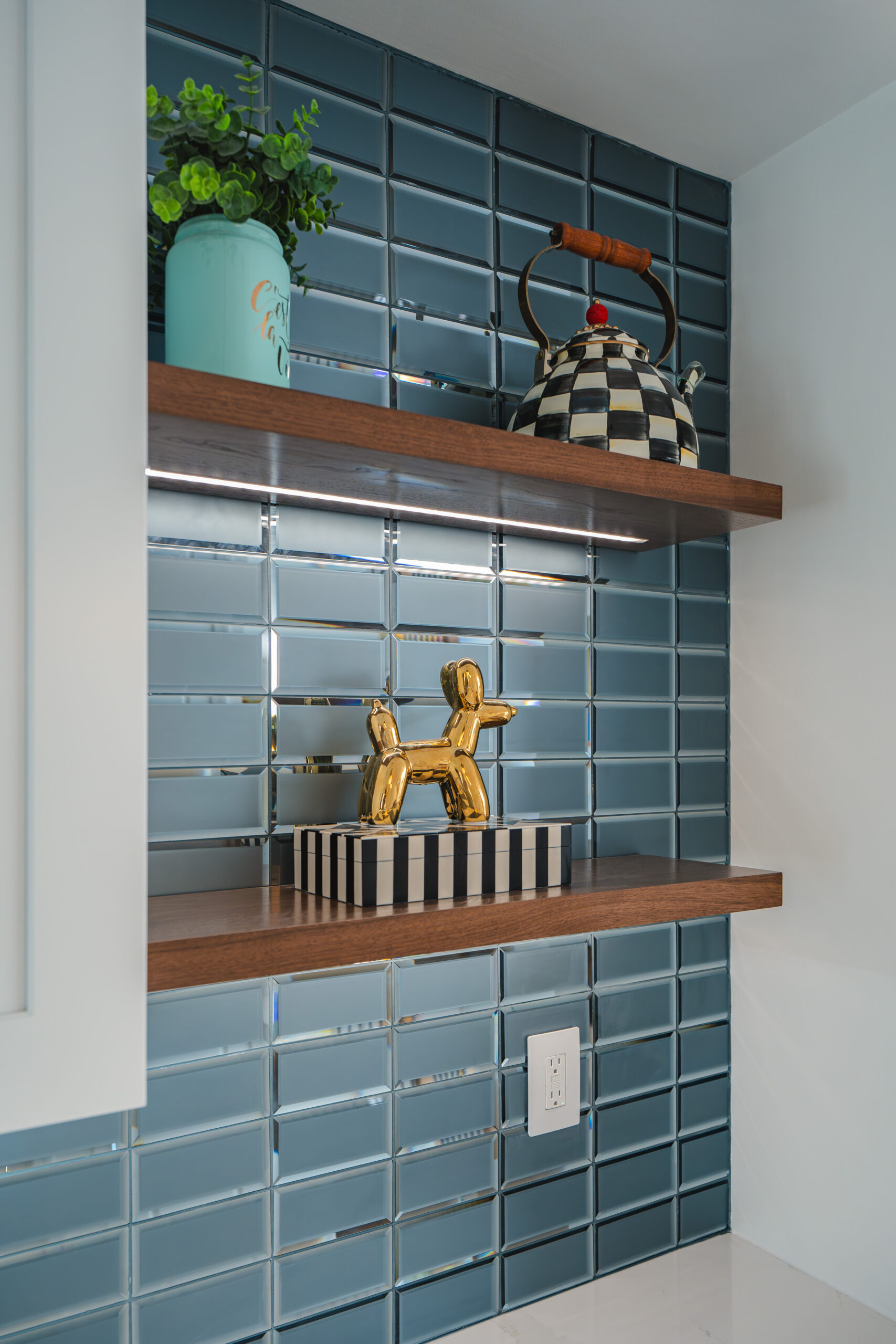 Dark wood floating shelves on a teal glass tile wall featuring a black and white checkered teapot and gold decor.