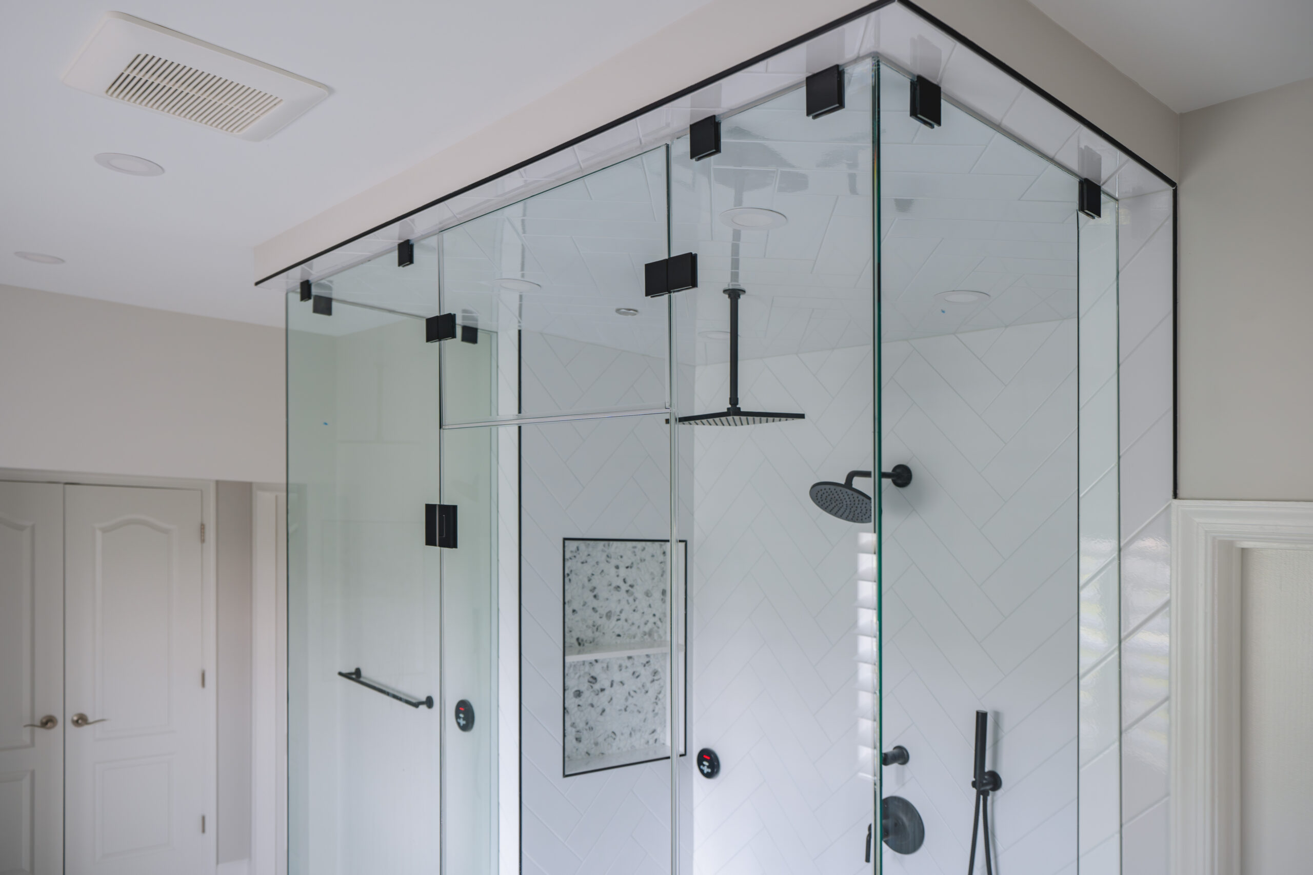 Looking up at the top of a frameless glass shower enclosure with matte black mounting clips and a ceiling-mounted rain shower head.