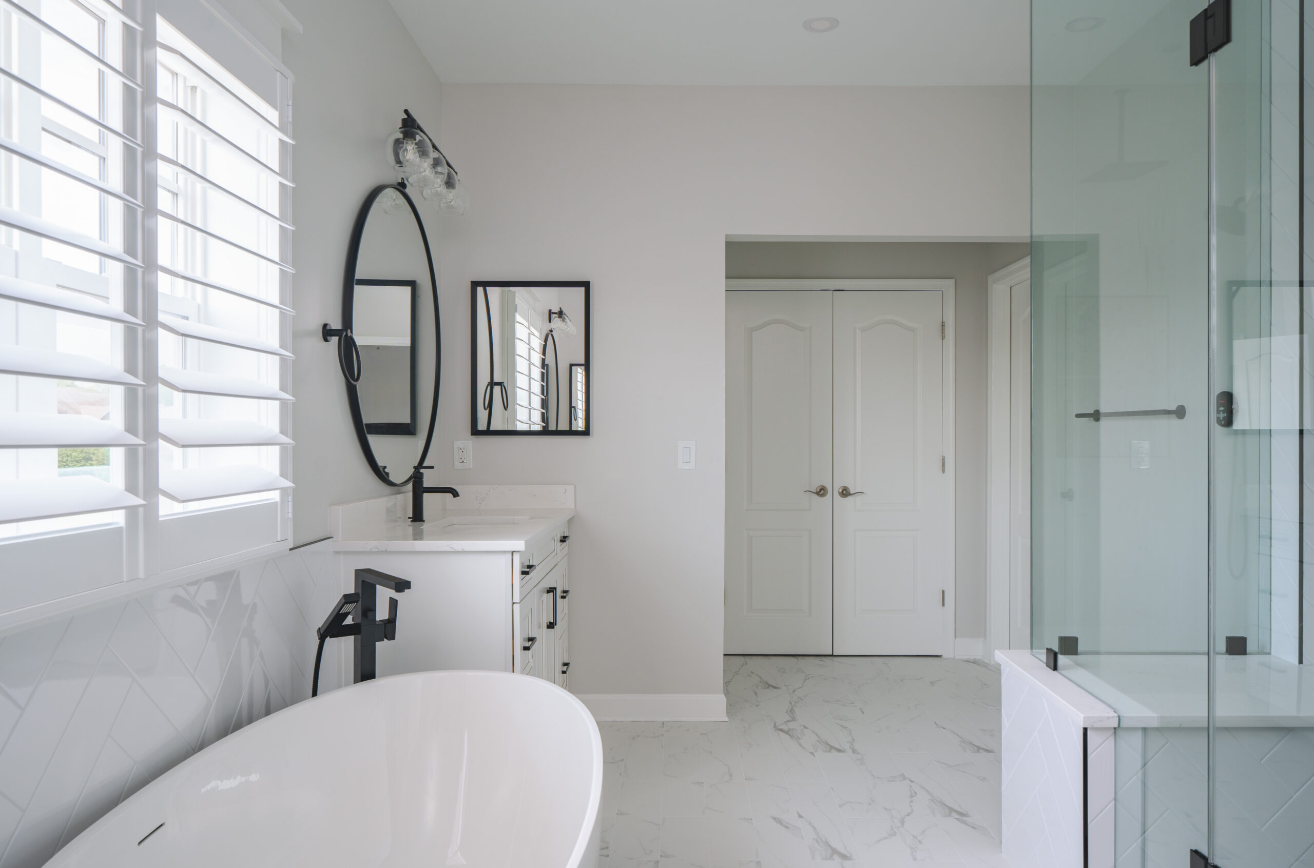 A white freestanding soaking tub positioned under large white window shutters next to a modern vanity.