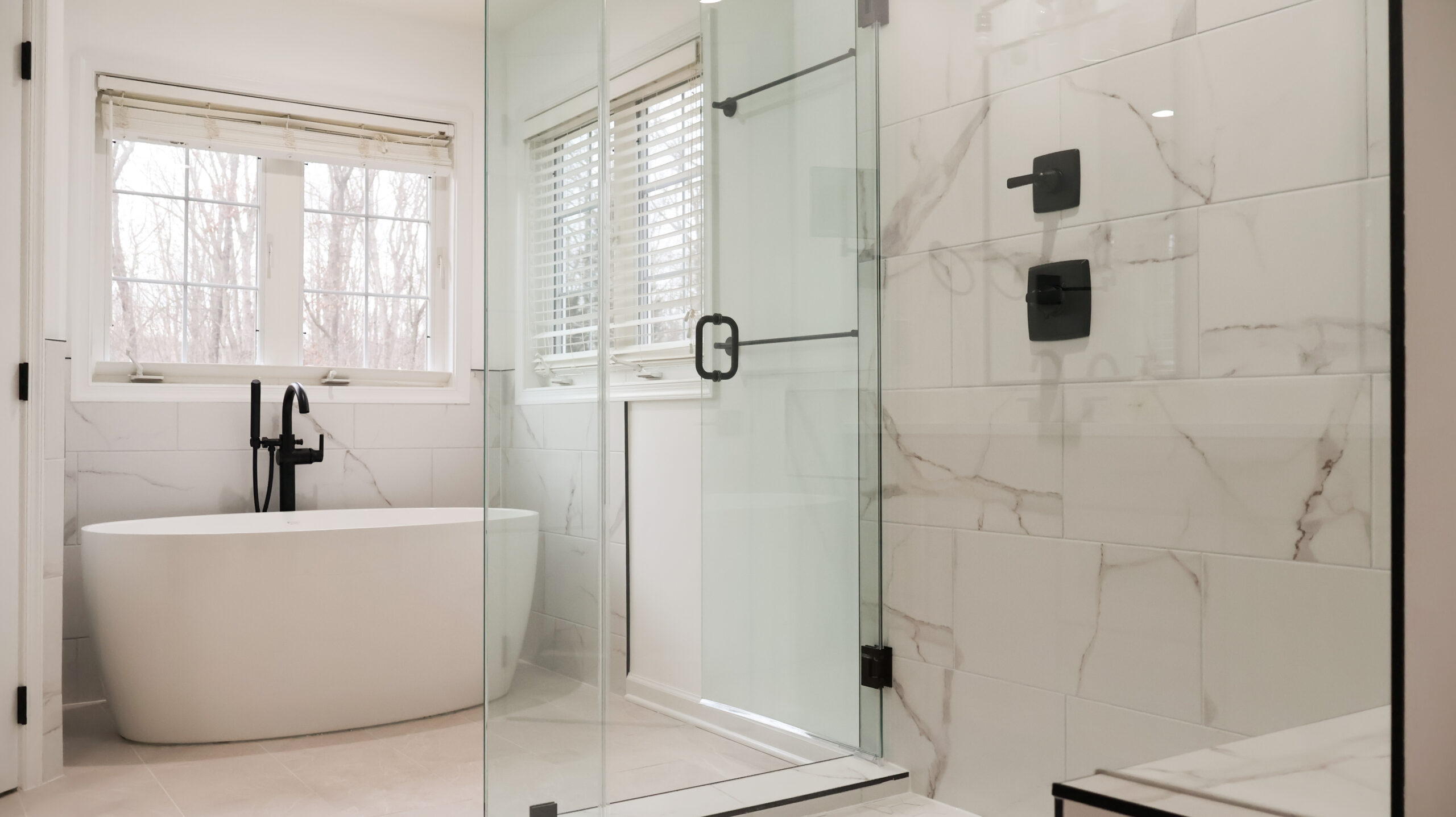 View from inside a walk in shower looking towards a freestanding tub, showcasing the glass door and black hardware.