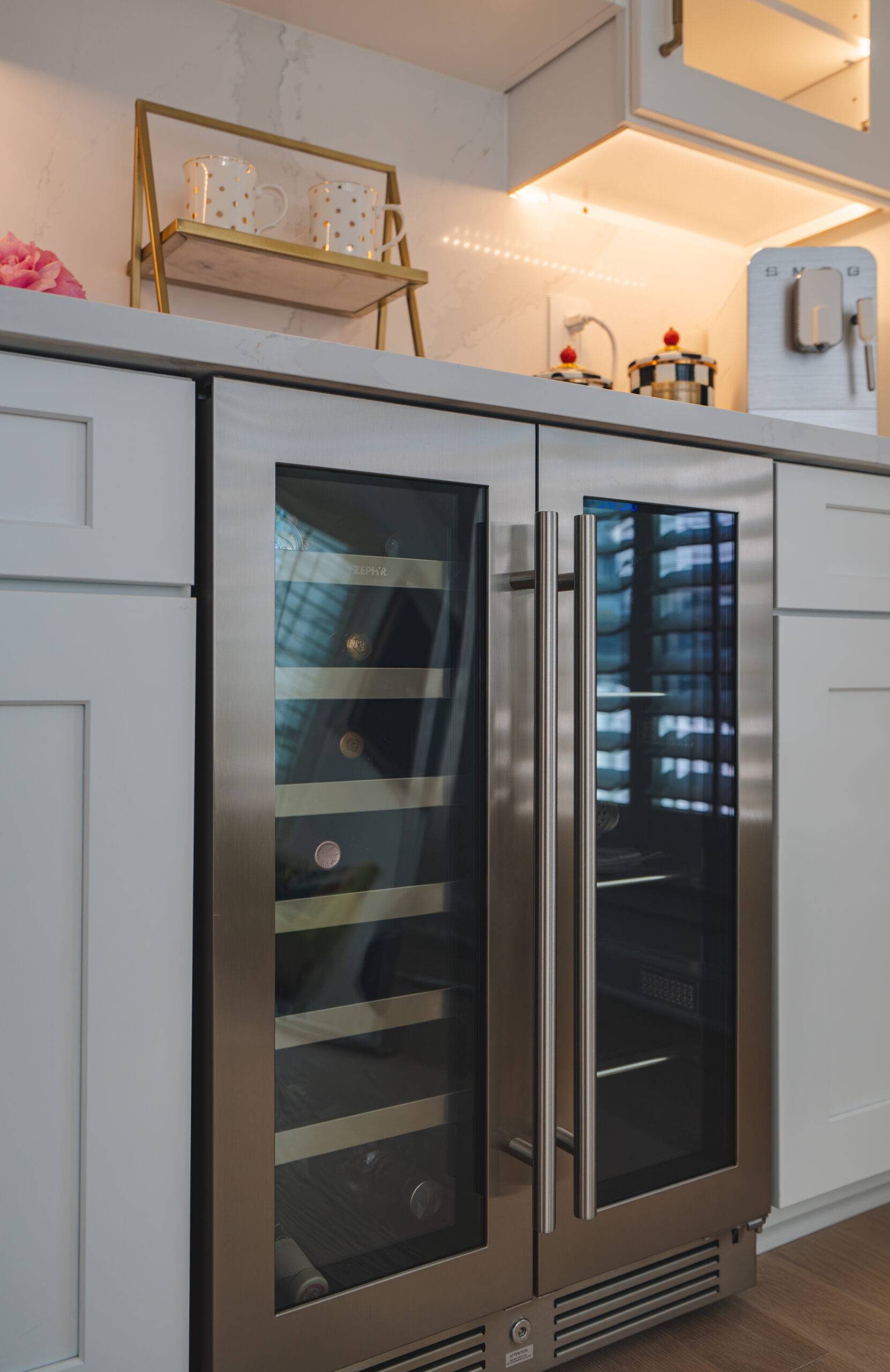 A stainless steel dual-zone Zephyr beverage and wine refrigerator built into white shaker-style cabinets.