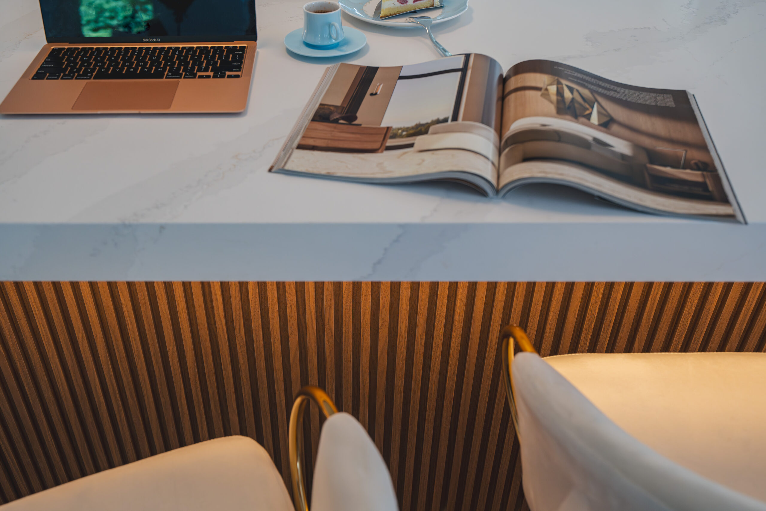 A lifestyle shot of a lit slatted wood kitchen island with white bar stools, a laptop, and an open magazine.