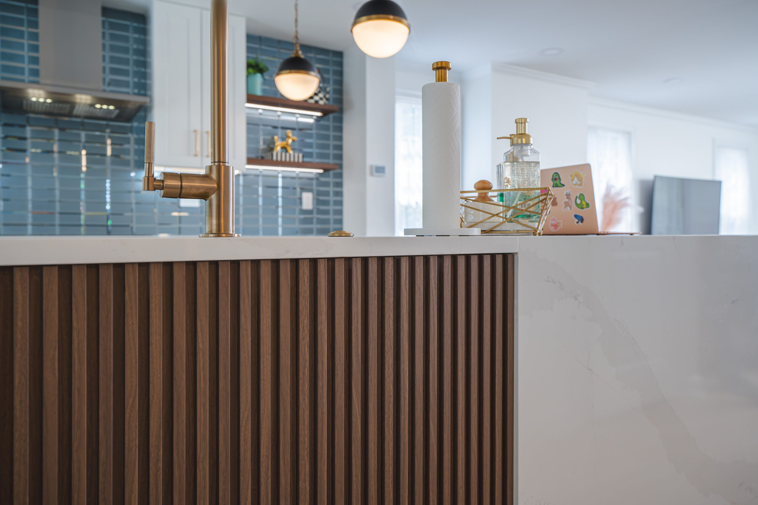 A close-up of a brushed gold gooseneck faucet installed on a white marble-look quartz island with fluted wood detail.