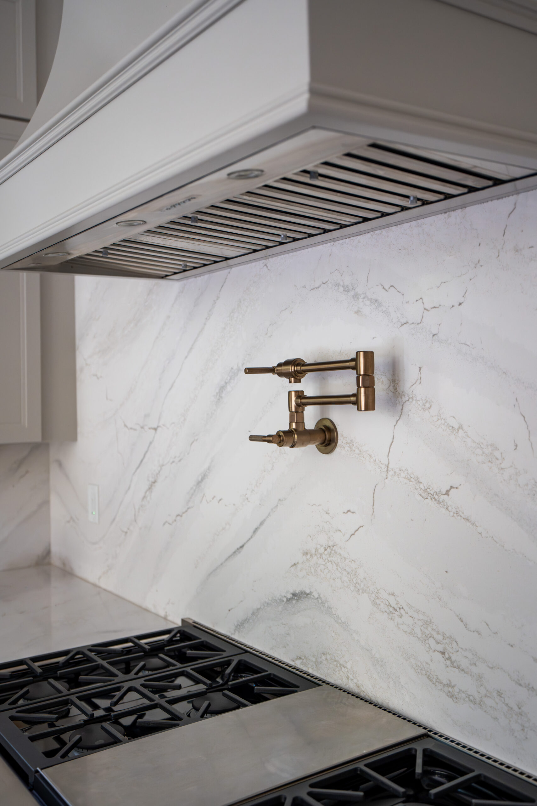 A sleek gold-toned pot filler faucet mounted on a marble backsplash above a gas range.