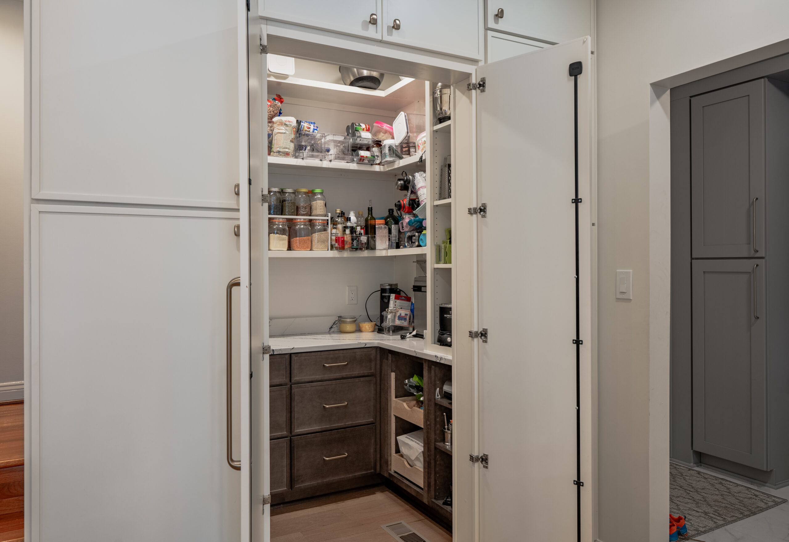 A walk-in kitchen pantry with white doors open, showing internal wood drawers, marble countertops, and organized shelving.