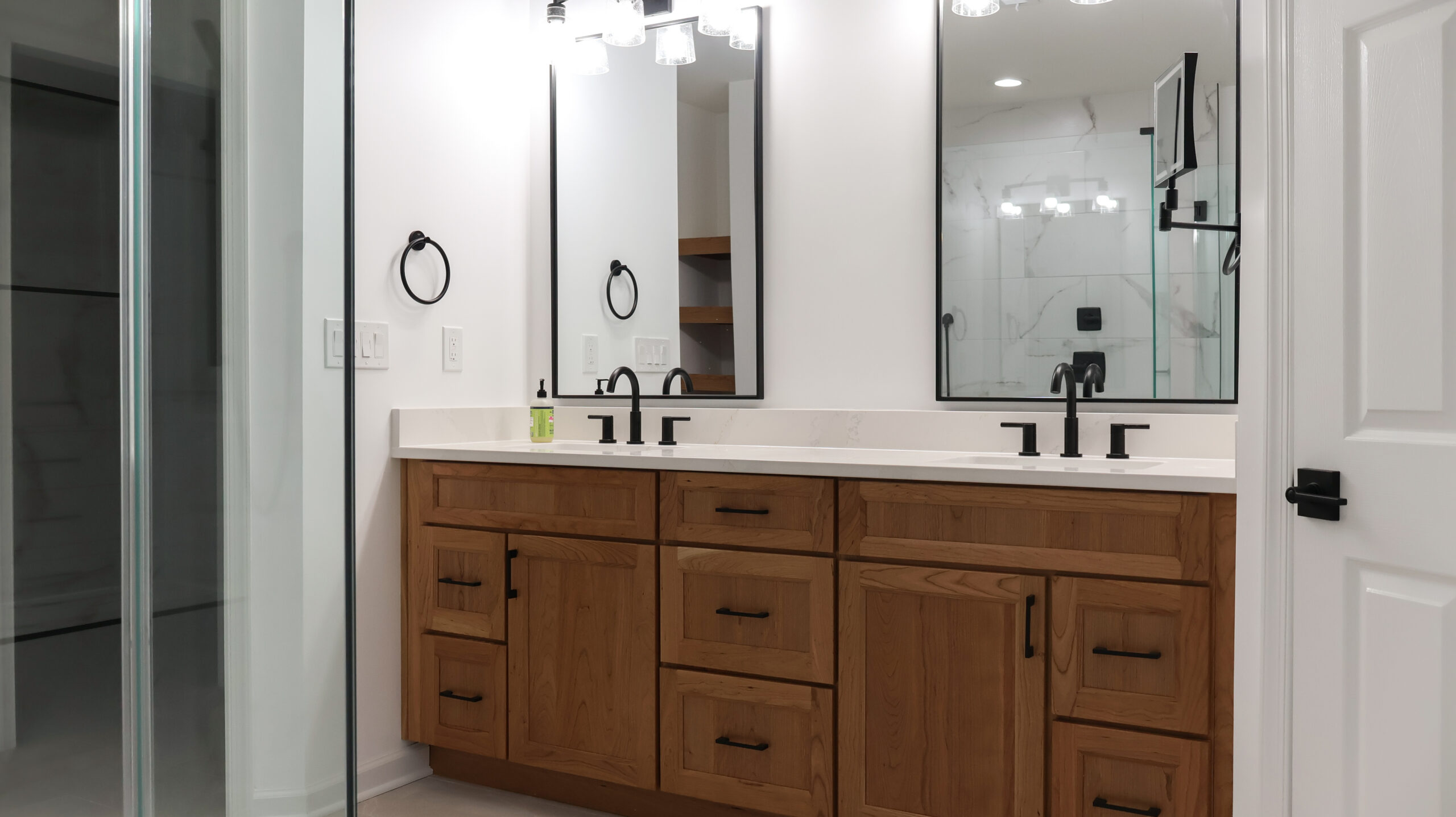 Custom light wood double bathroom vanity with white quartz countertops, black faucets, and black framed rectangular mirrors.