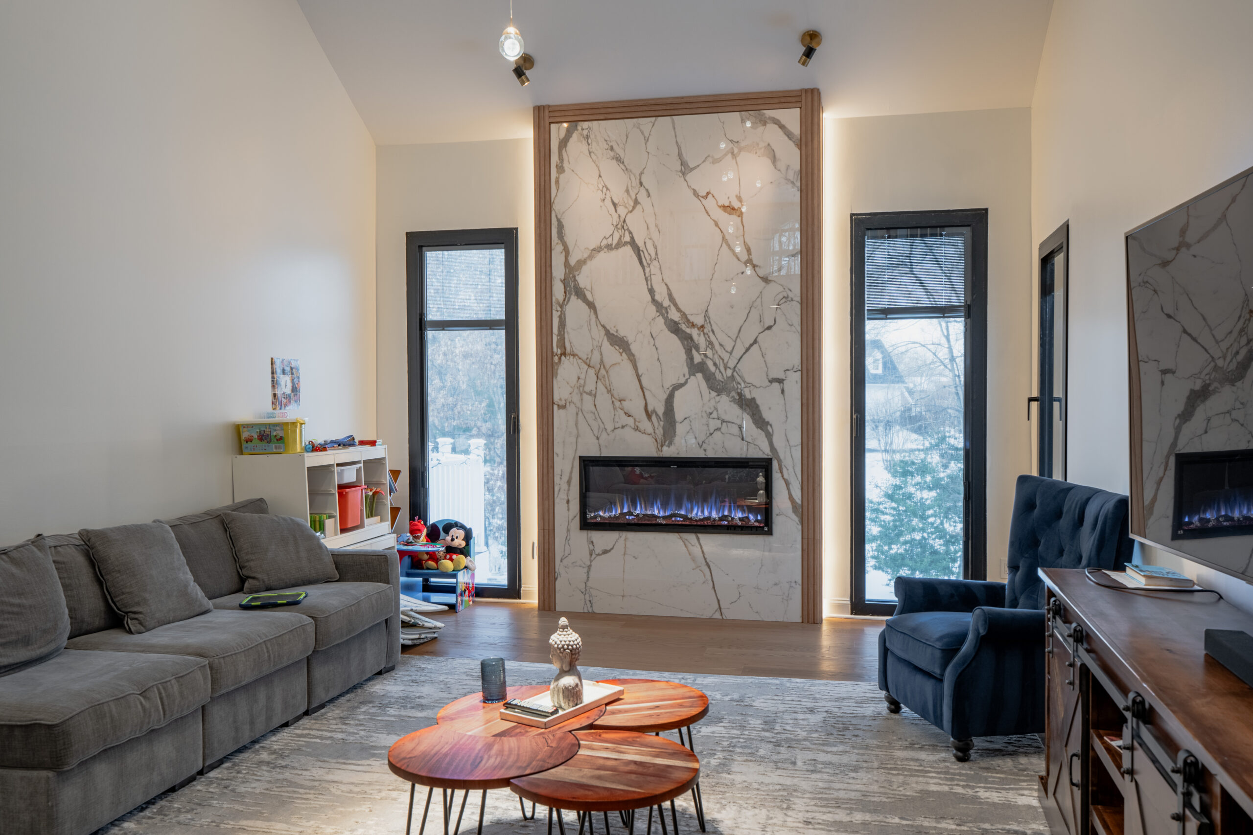 A wide view of a renovated living room featuring a marble fireplace, modern furniture, and large black-framed windows.
