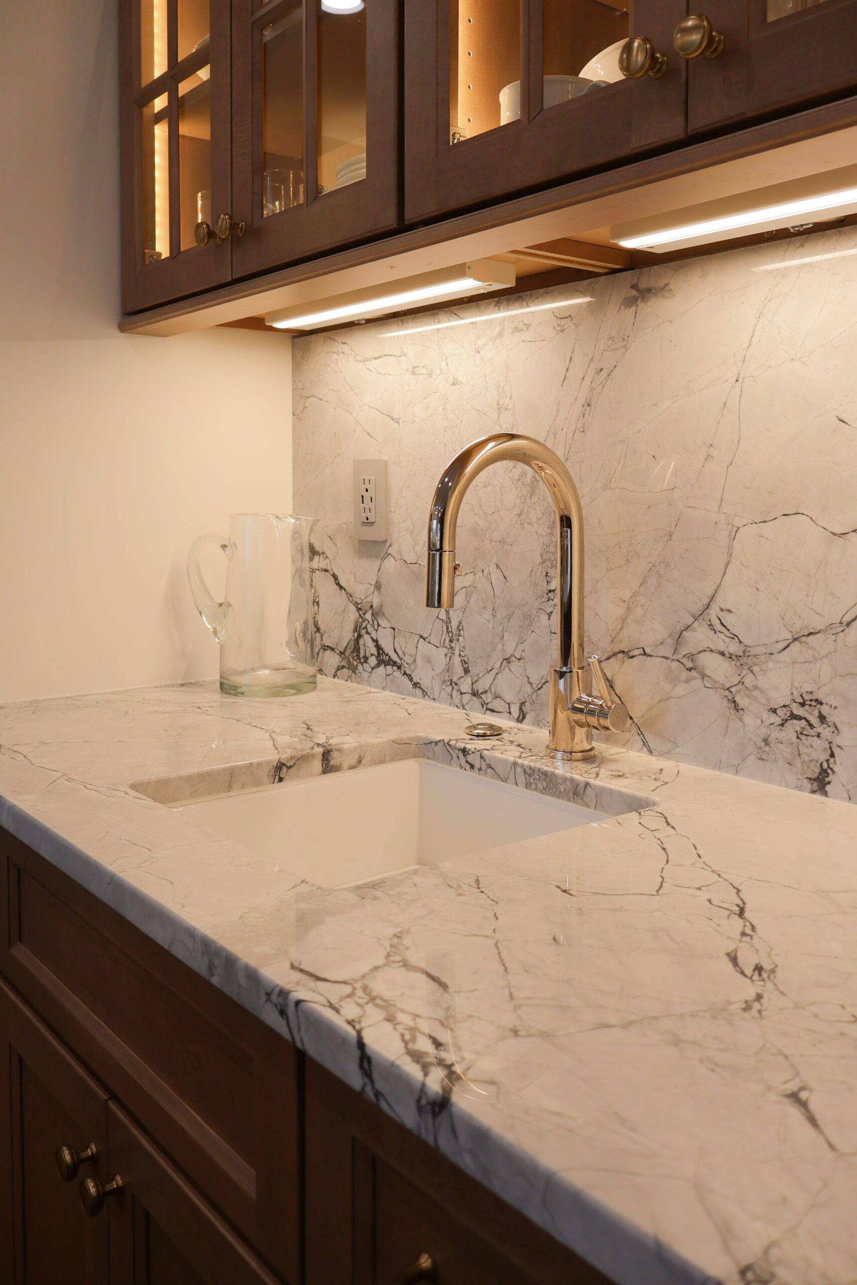 Home beverage center and wet bar in Georgetown DC featuring dark wood cabinets, a gold faucet, and lit glass-front upper cabinets.