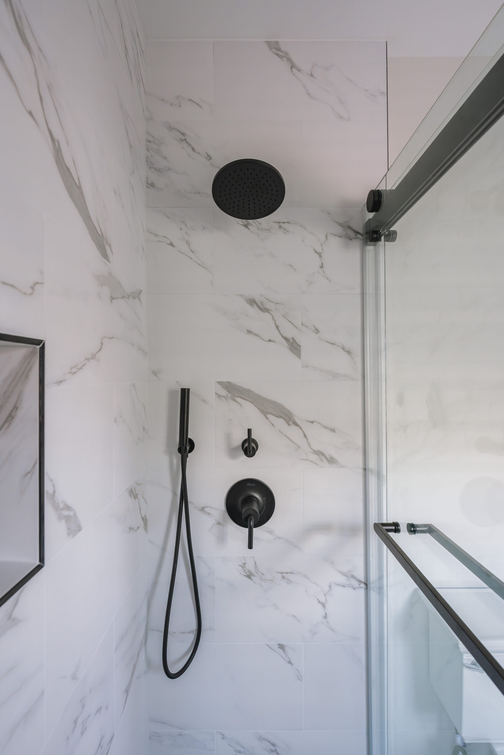 A shower interior with a large matte black rain shower head, handheld sprayer, and matching control valves on marble-veined walls.