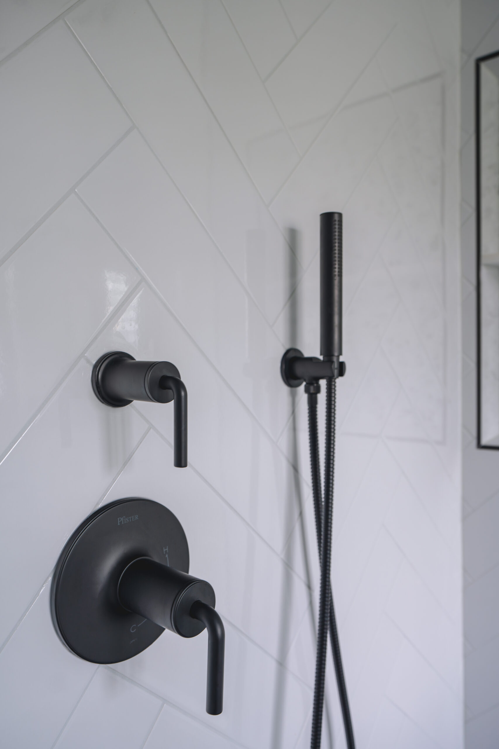 A close-up of matte black Pfister shower handles and a handheld sprayer mounted on white herringbone wall tile.