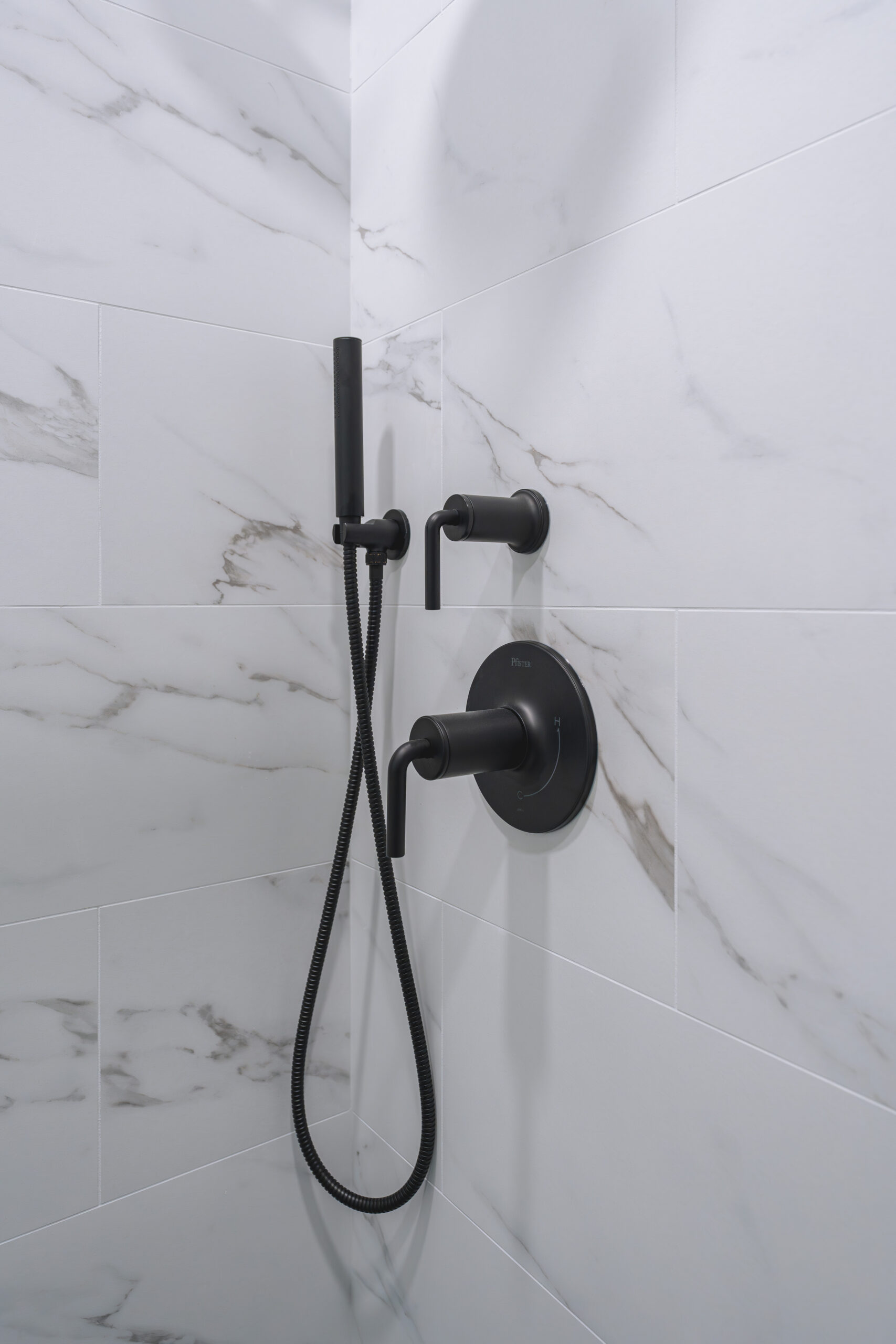 Matte black shower control handles and a handheld wand sprayer mounted on large format marble tiles.