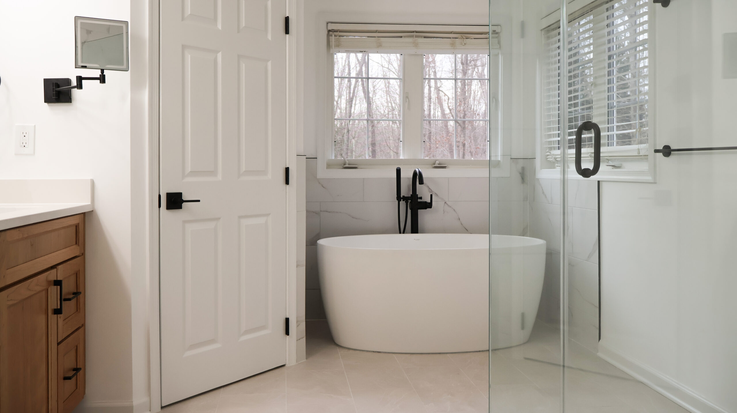 Modern bathroom featuring a white freestanding tub, matte black faucet, and light wood double vanity with marble look tile walls.