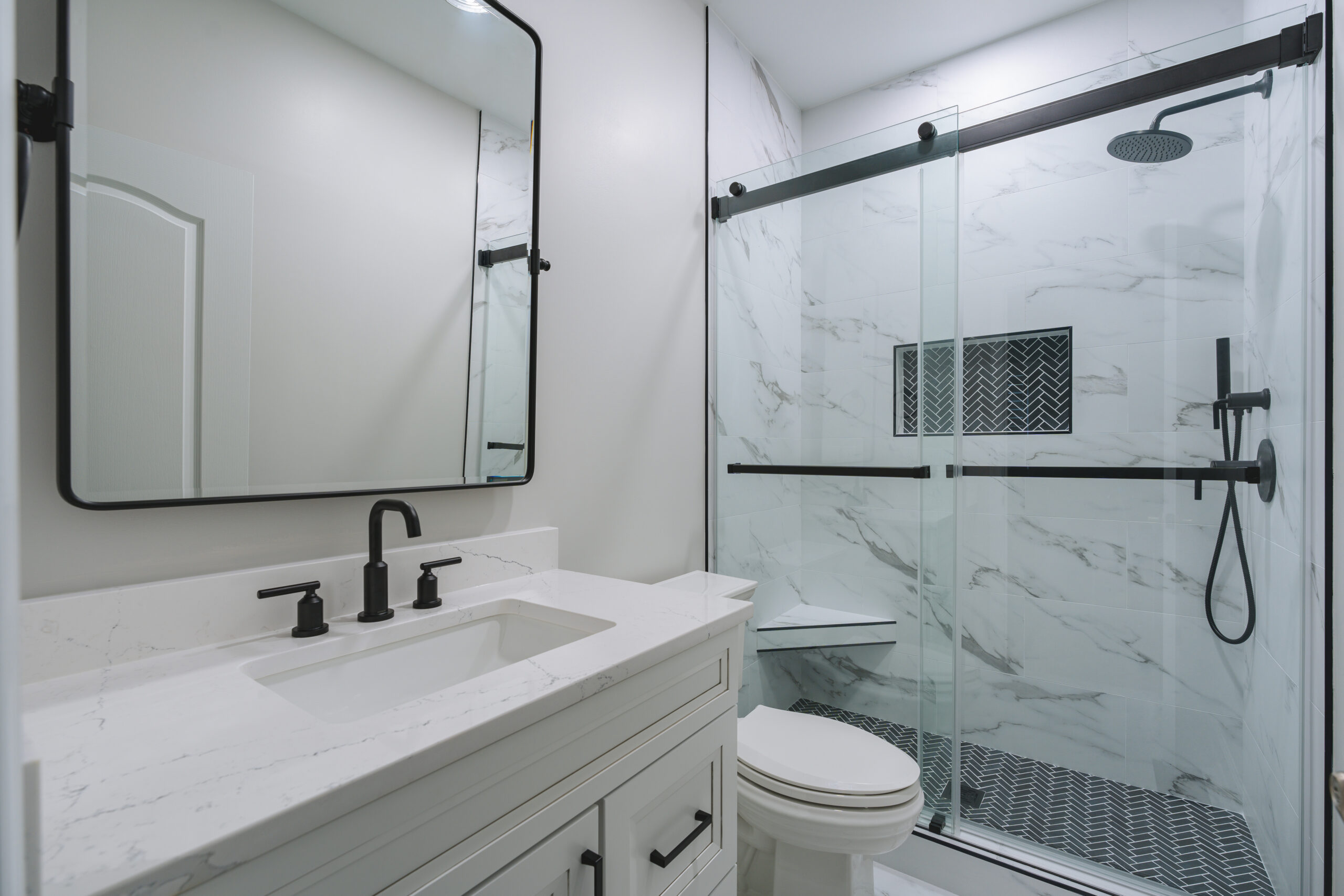 A modern bathroom view including a white vanity, toilet, and a sliding glass shower door with marble-patterned walls.