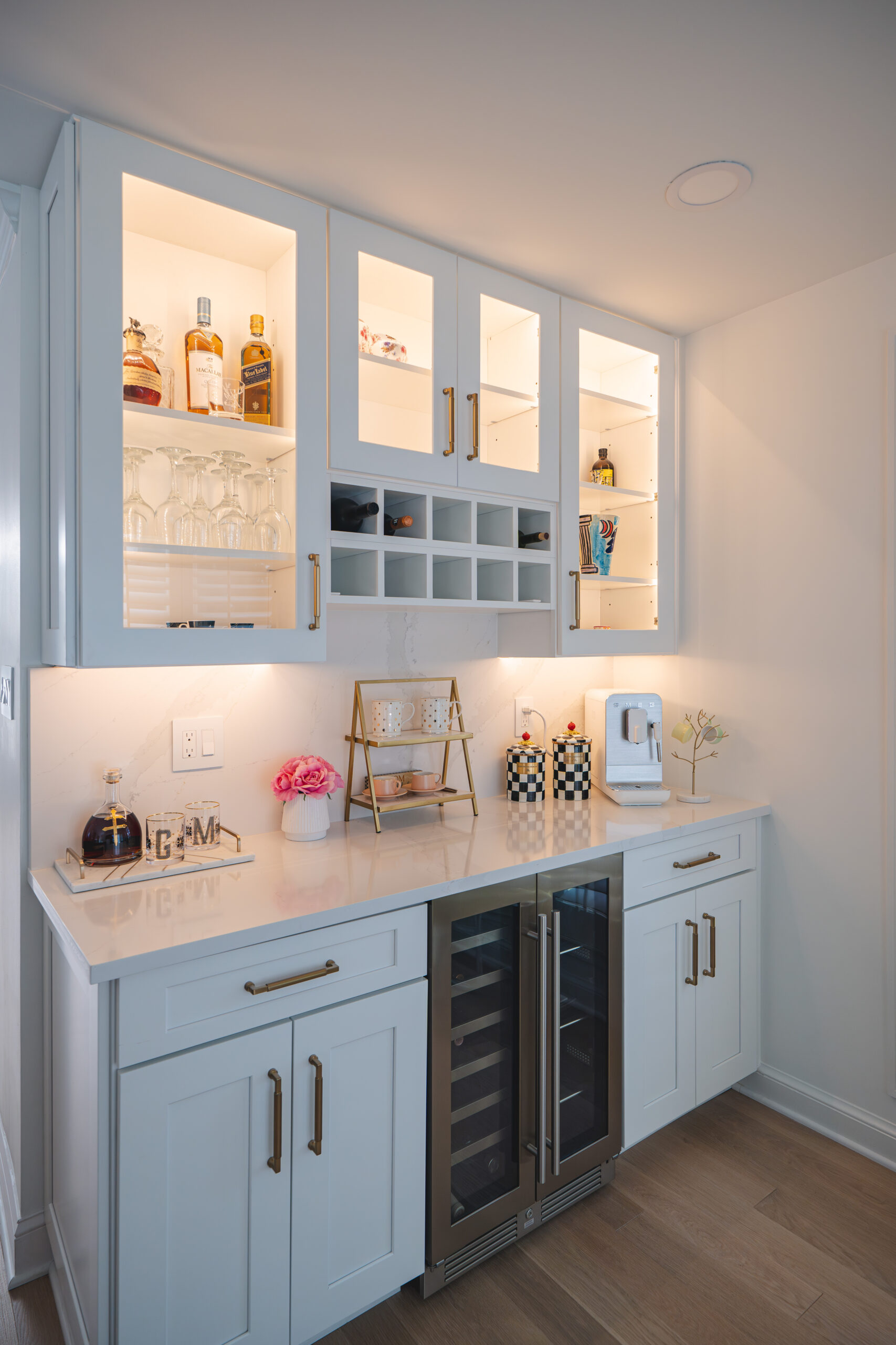 A complete view of a modern dry bar with a dual-zone wine fridge, lit upper cabinets, and gold hardware.