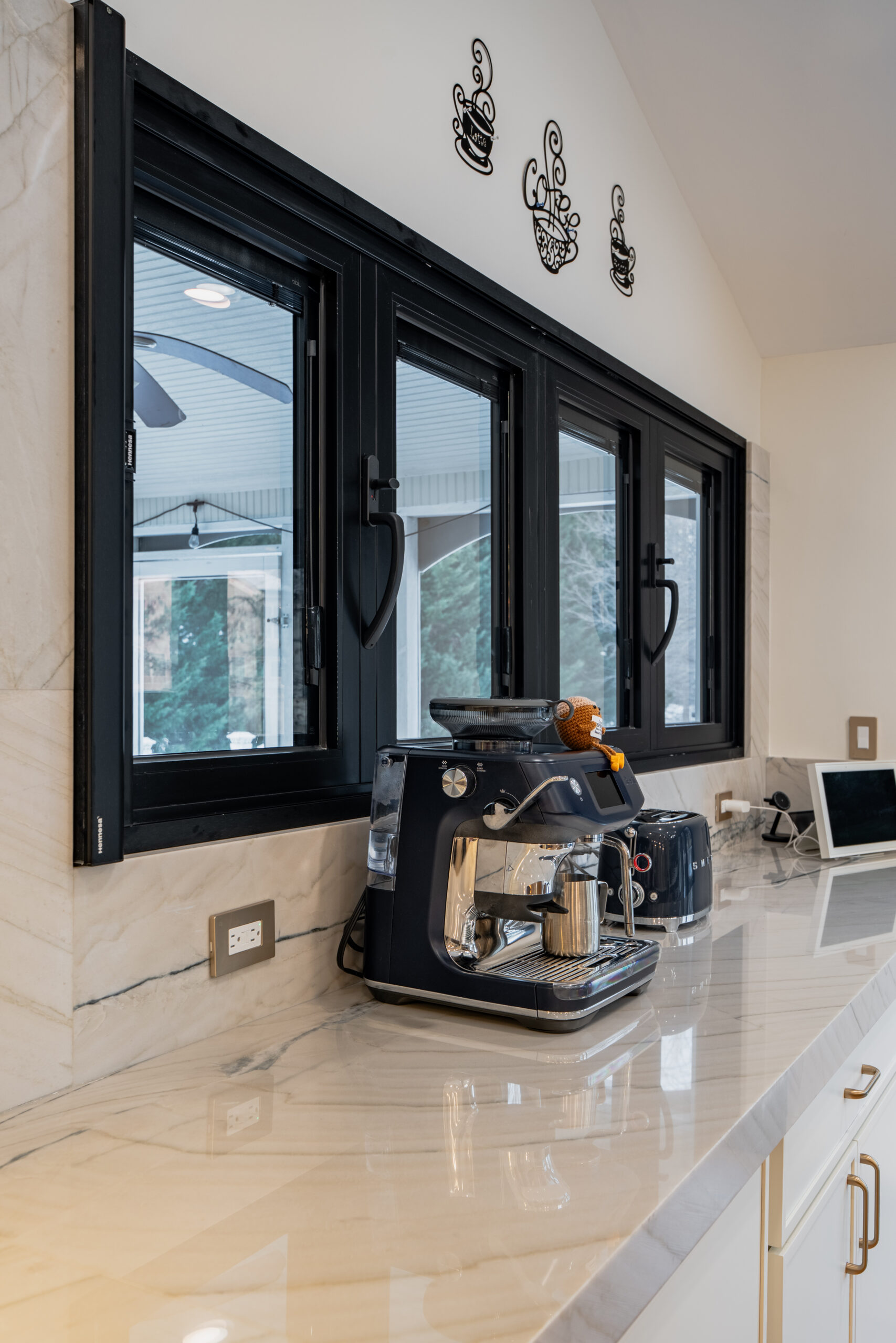 A high-end espresso machine and accessories on a marble kitchen countertop in front of black-framed windows.