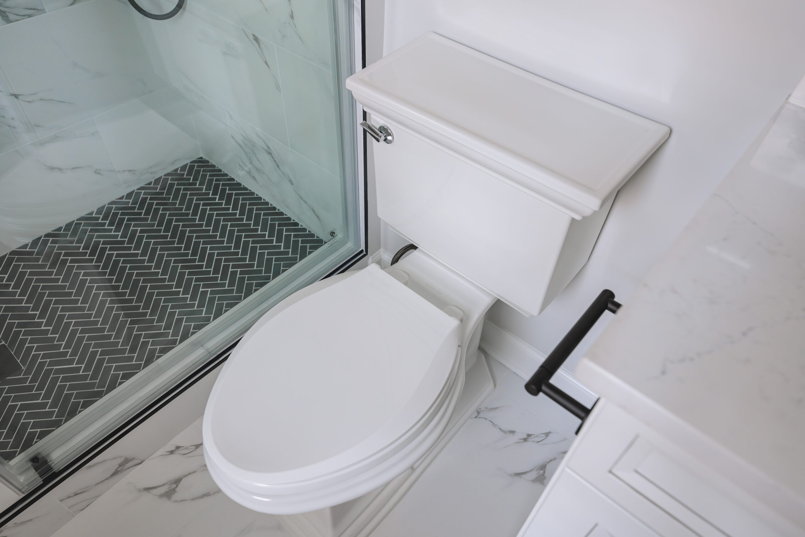 A top-down view of a white toilet on marble-veined floor tiles next to a glass shower with black herringbone floor.