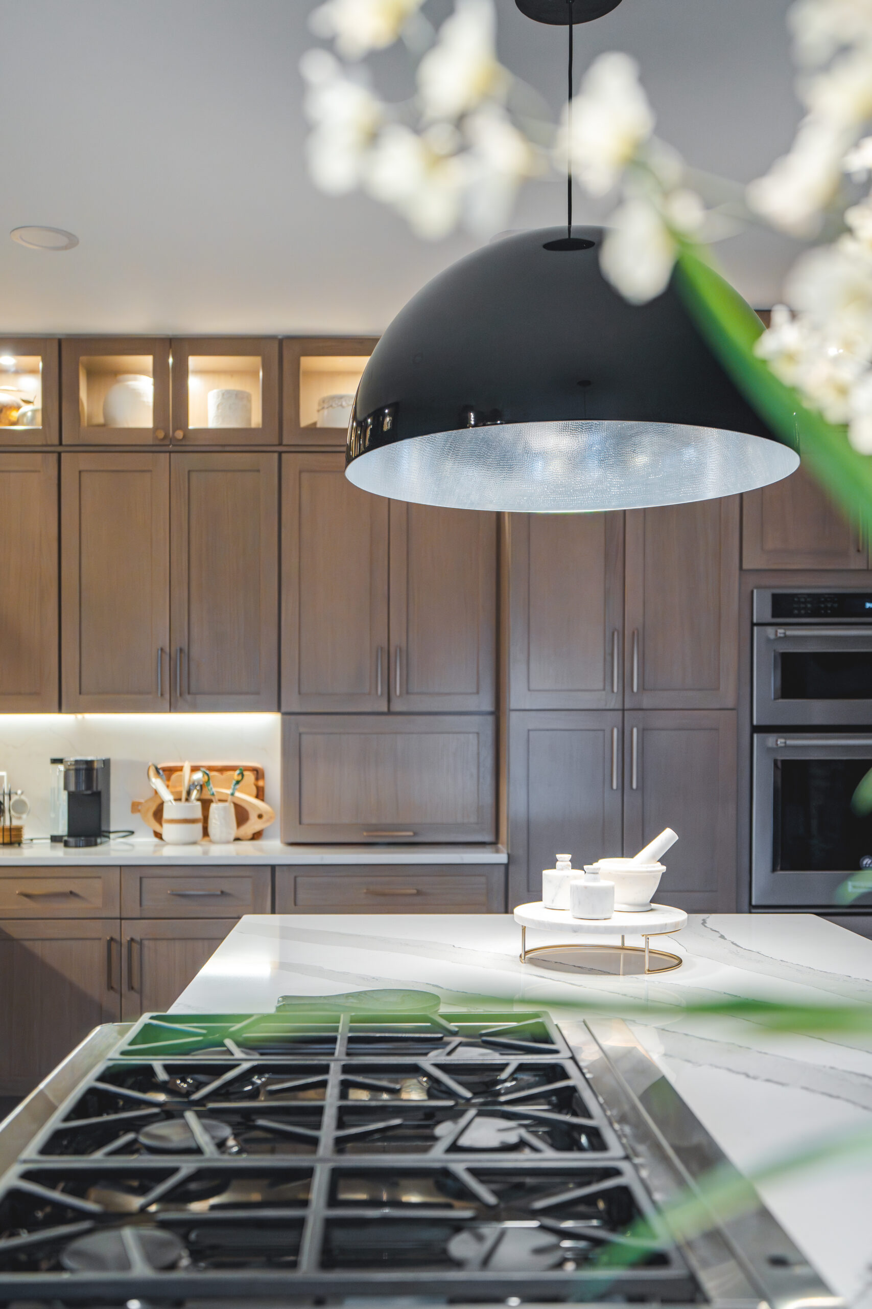 A creative shot showing white flowers in the foreground and a large black dome pendant light over a marble quartz island.