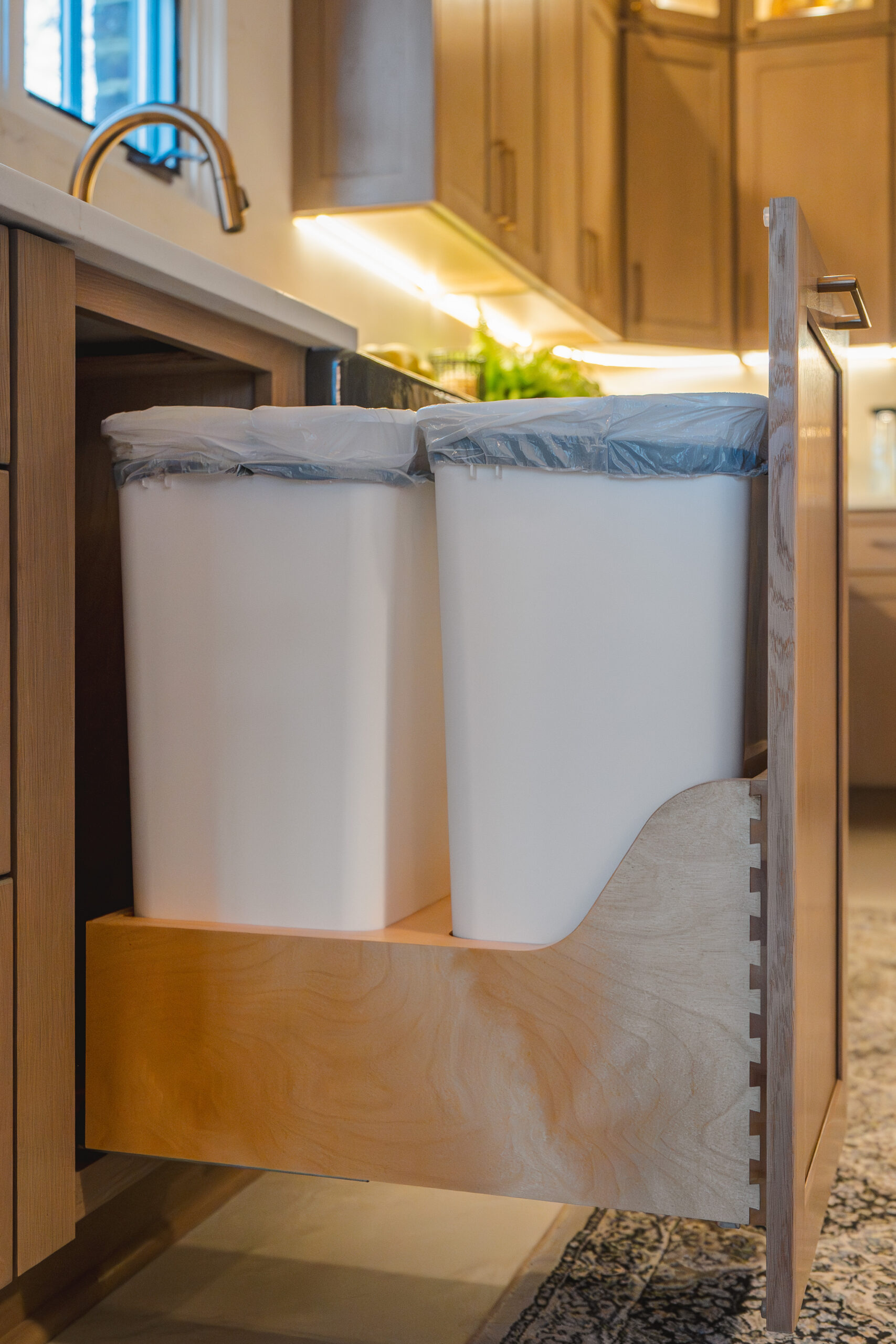 A pull-out wooden cabinet drawer with two white trash cans integrated into the kitchen's perimeter cabinetry.