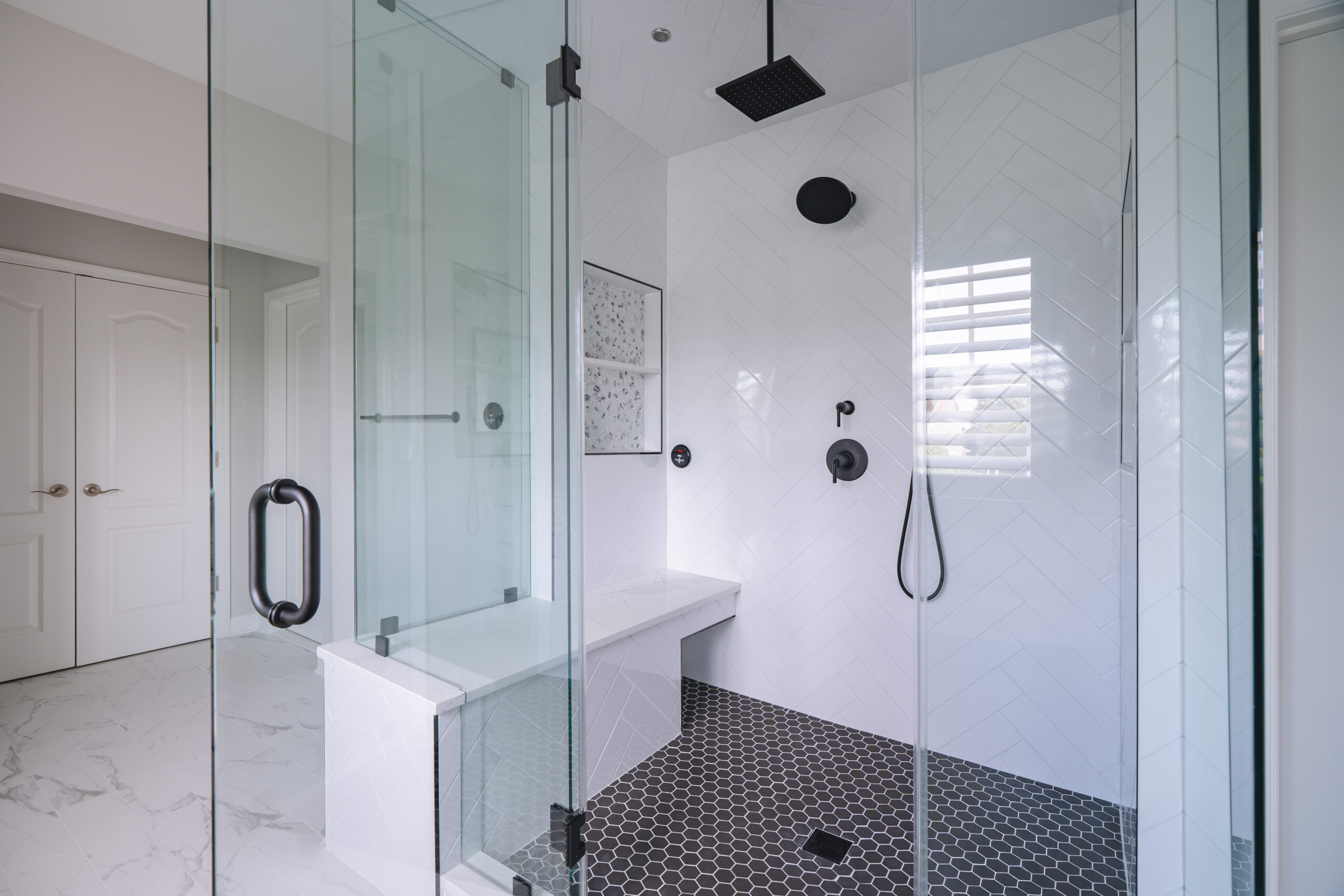 The interior of a shower showing a white marble bench, white herringbone walls, and black hexagon floor tiles.