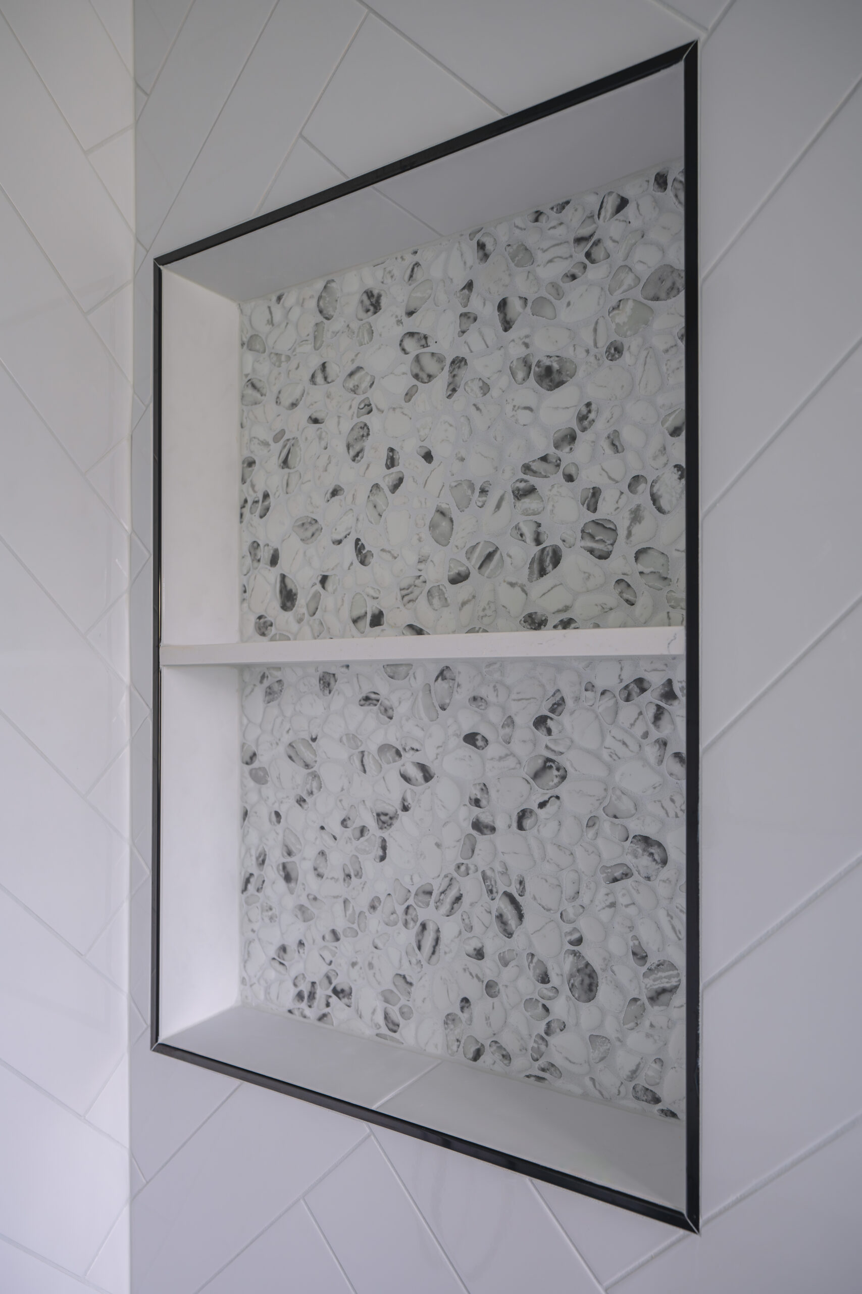A recessed wall niche in a shower featuring a gray and white pebble tile mosaic backing and black trim.