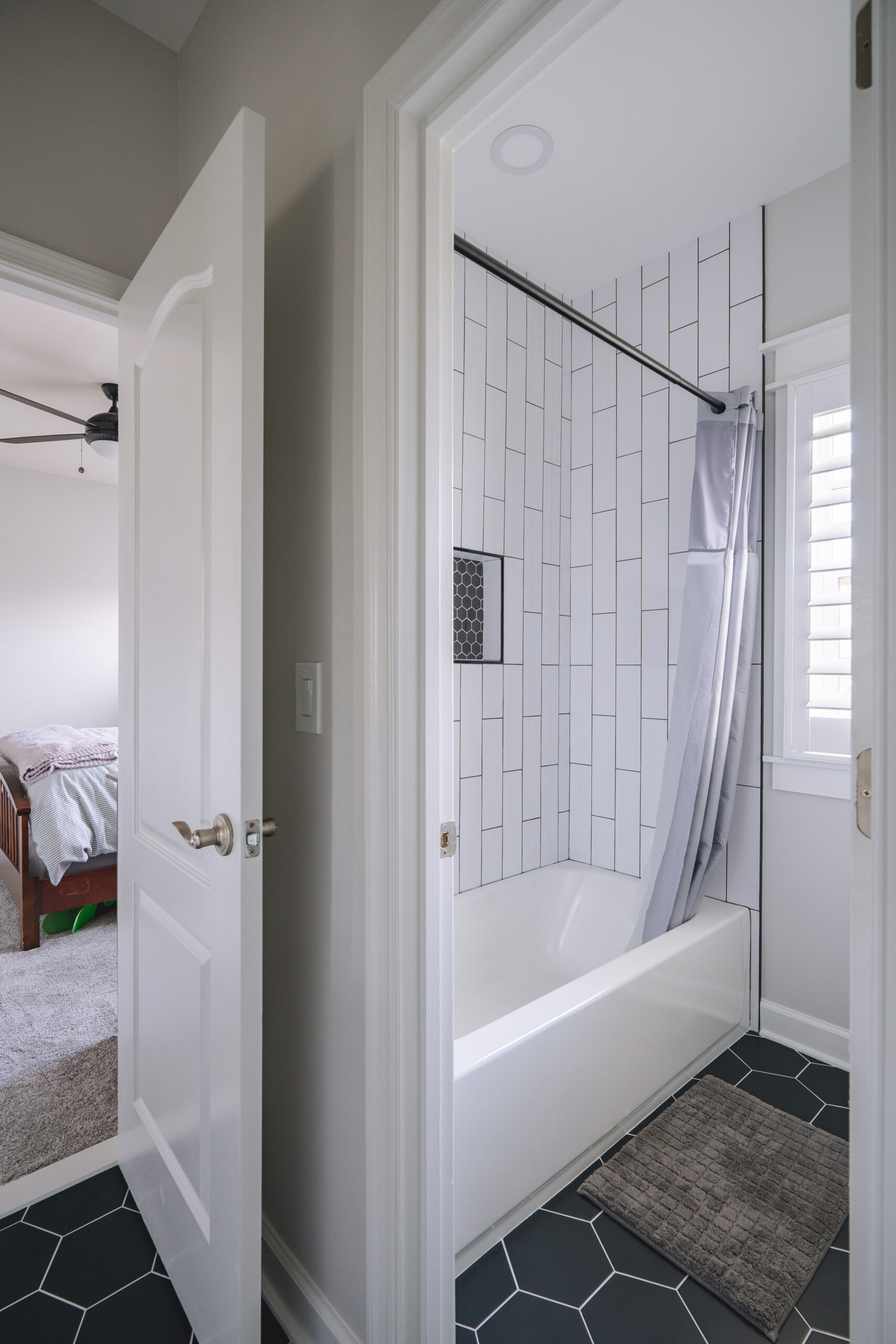 A view into a bathroom featuring a tub-shower combo with white vertical subway tile and black hexagon floor mosaics.