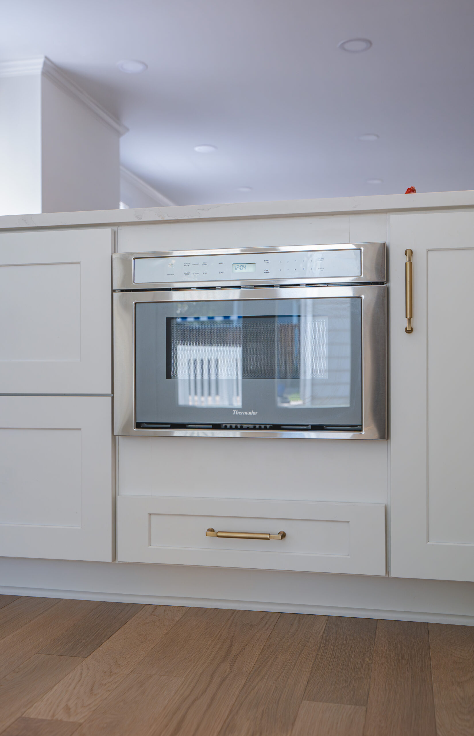 A stainless steel Thermador microwave drawer built into white shaker-style cabinetry with gold pulls.