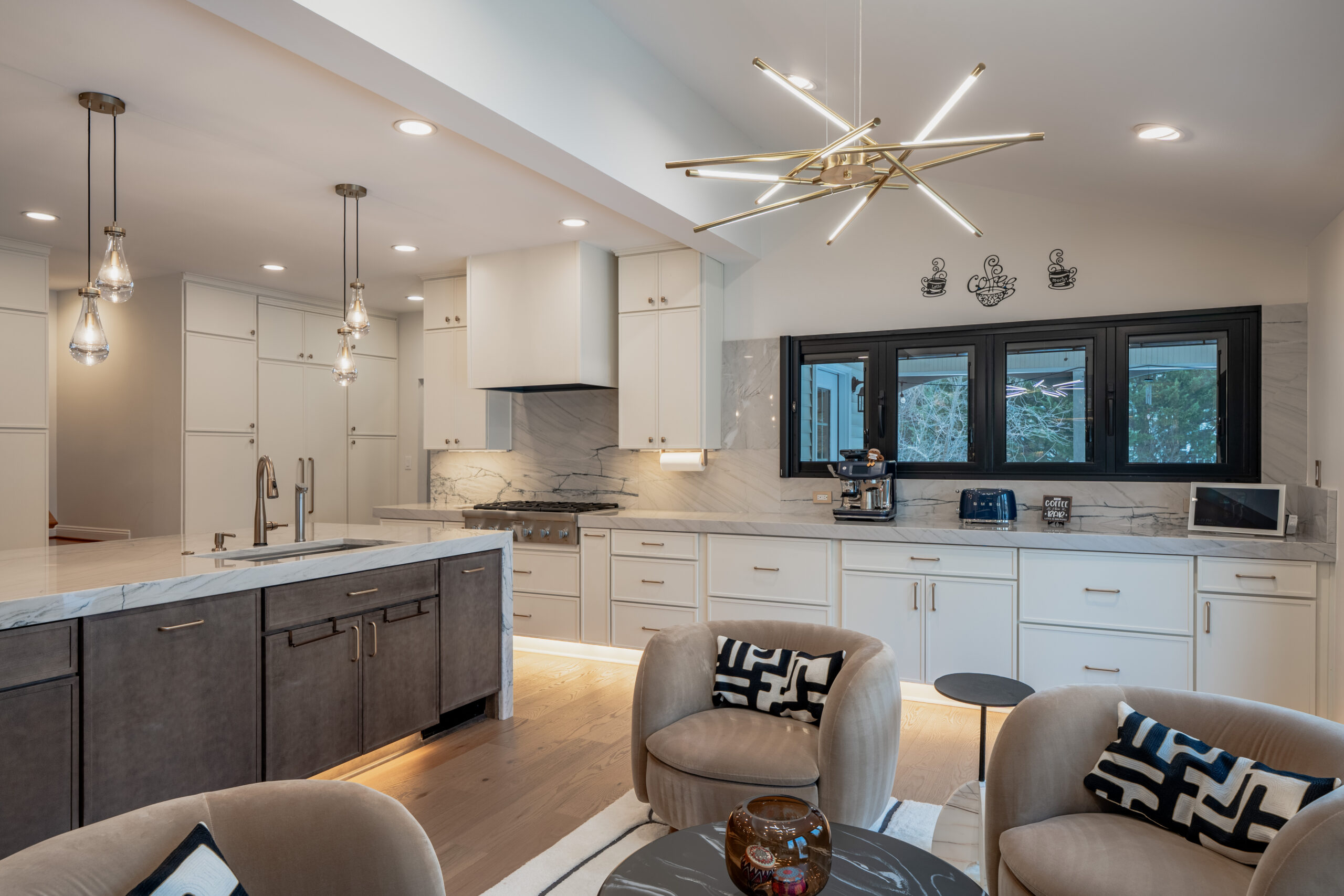 A side view of a large kitchen island with gray upholstered stools and a modern gold chandelier in the living space.
