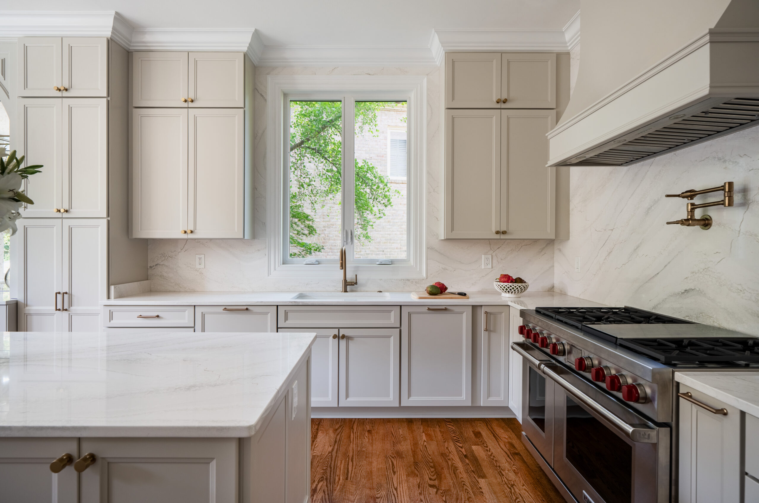 A modern kitchen sink with a gold faucet set into a marble countertop under a large window with a garden view.