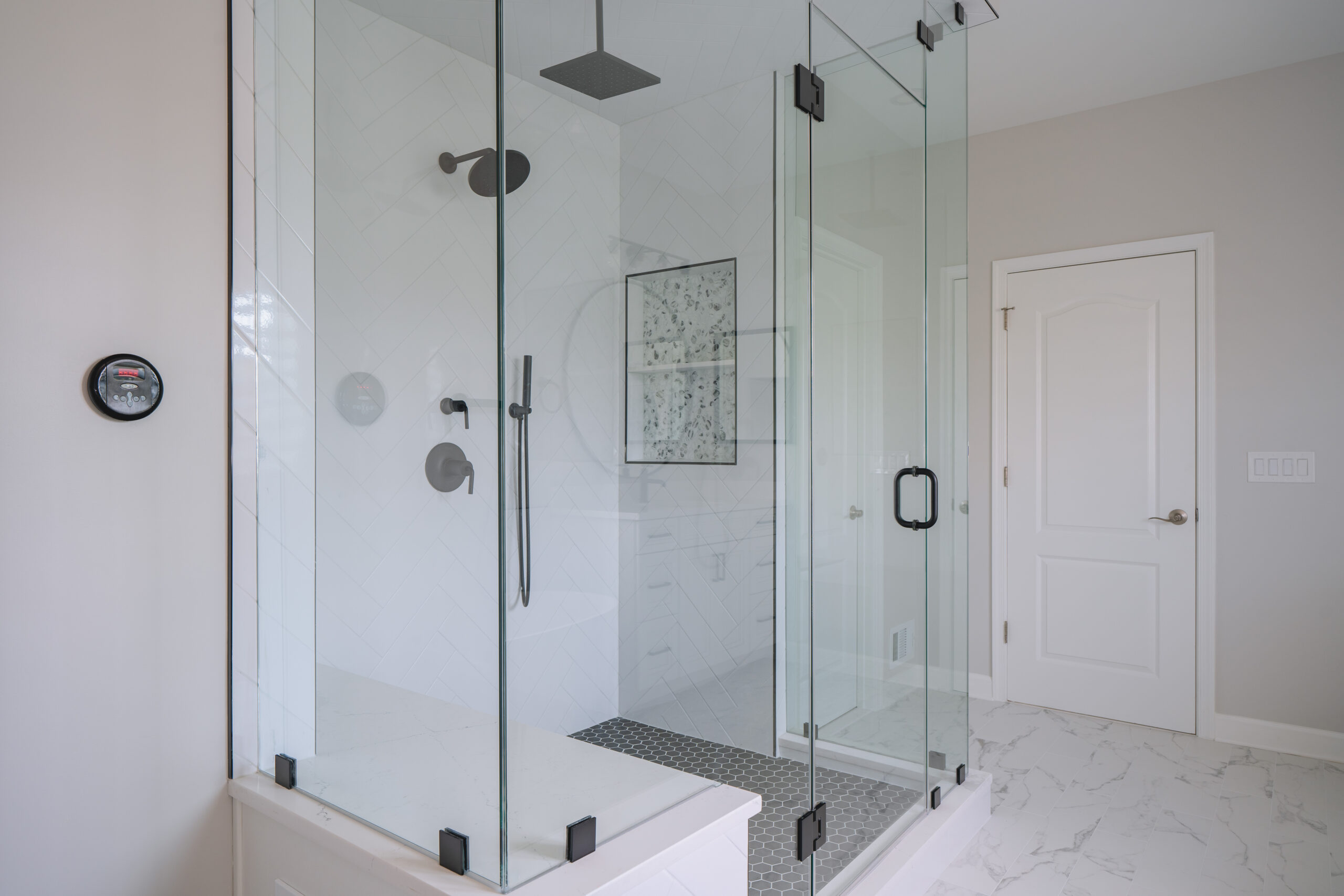A frameless glass walk-in shower with white herringbone wall tiles and black rain shower heads.