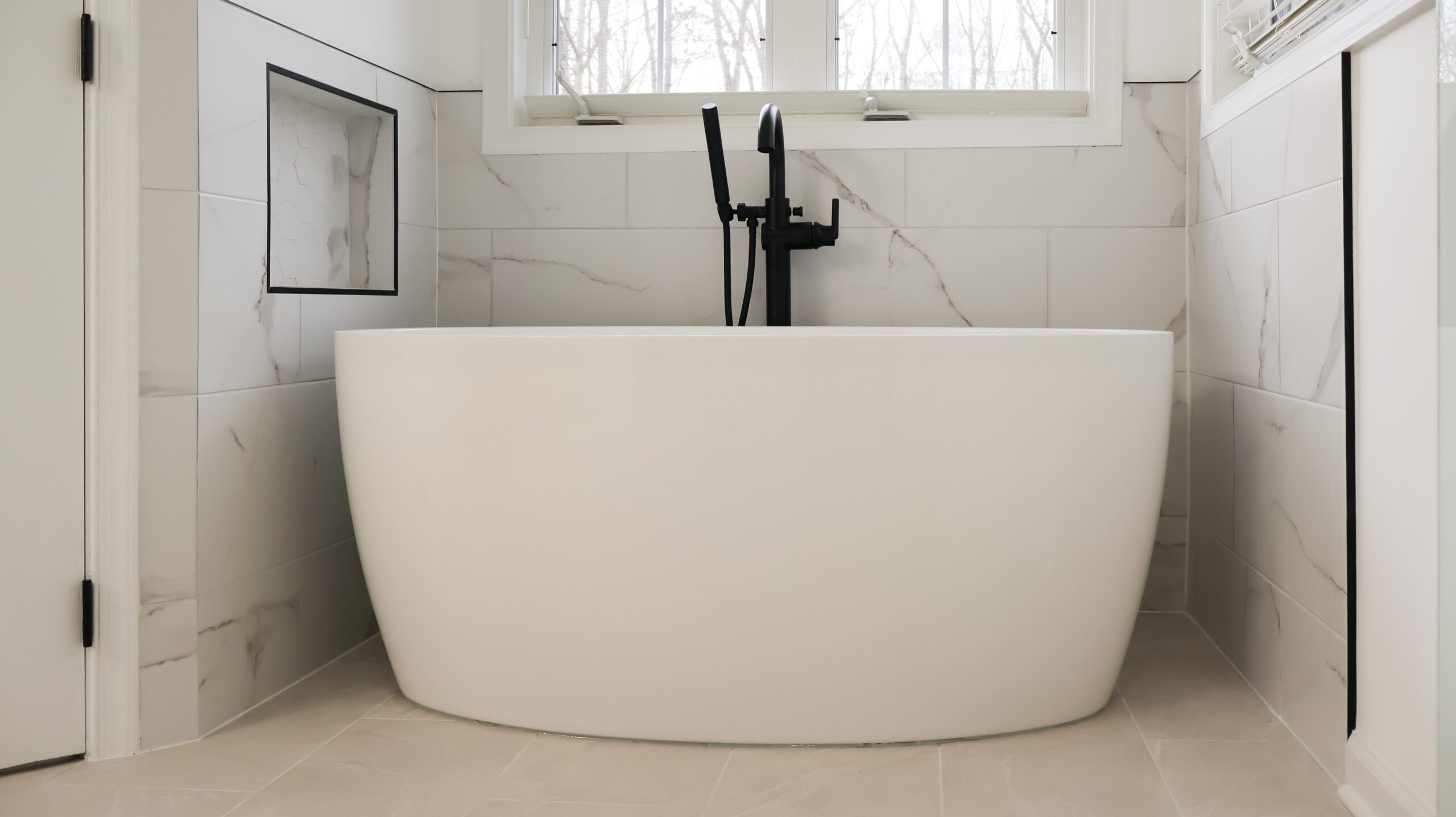 Close up of a contemporary white freestanding soaking tub with a floor mounted matte black faucet and marble patterned wall tiles.