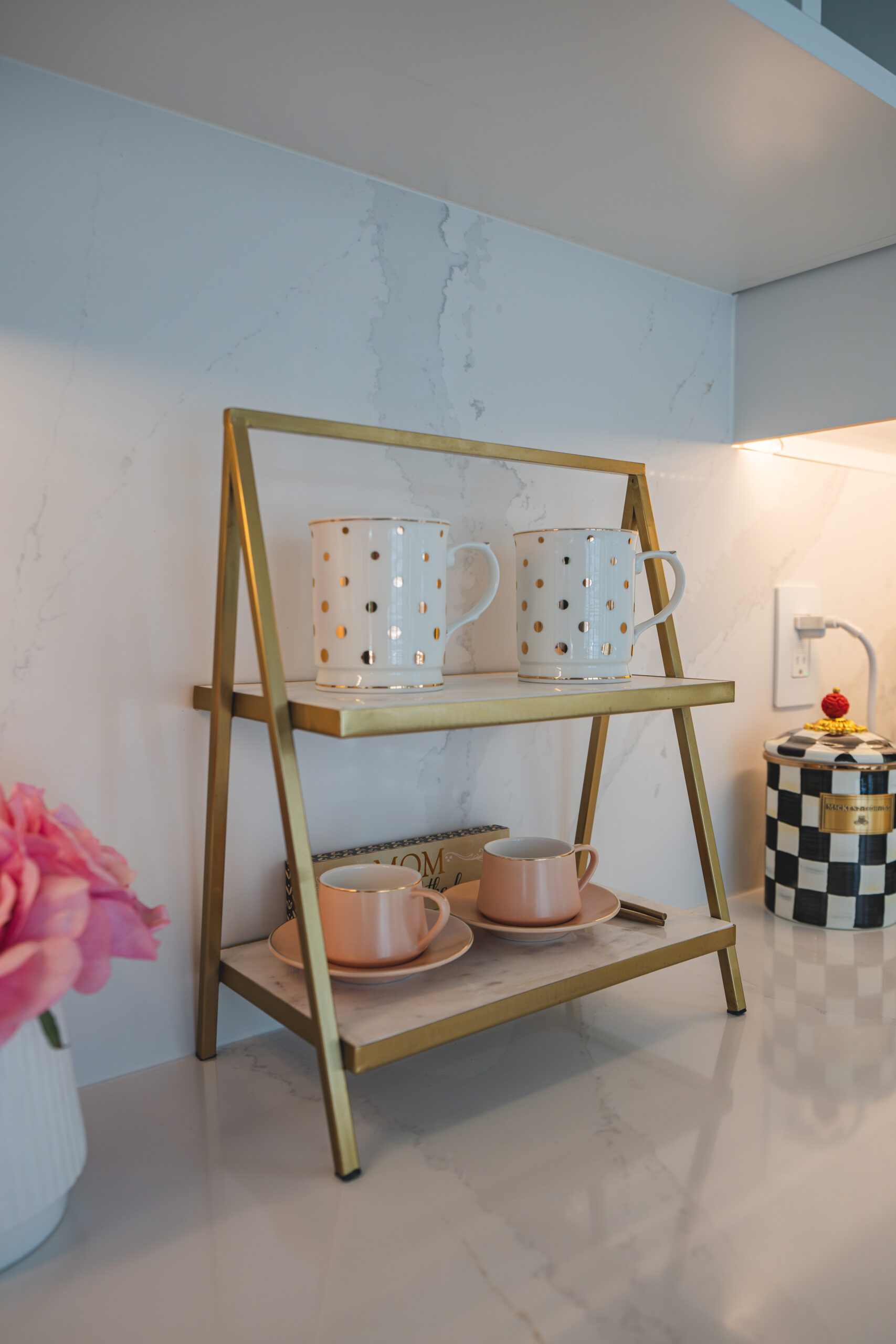 A gold tiered display rack with polka dot mugs and pink teacups on a white quartz countertop.
