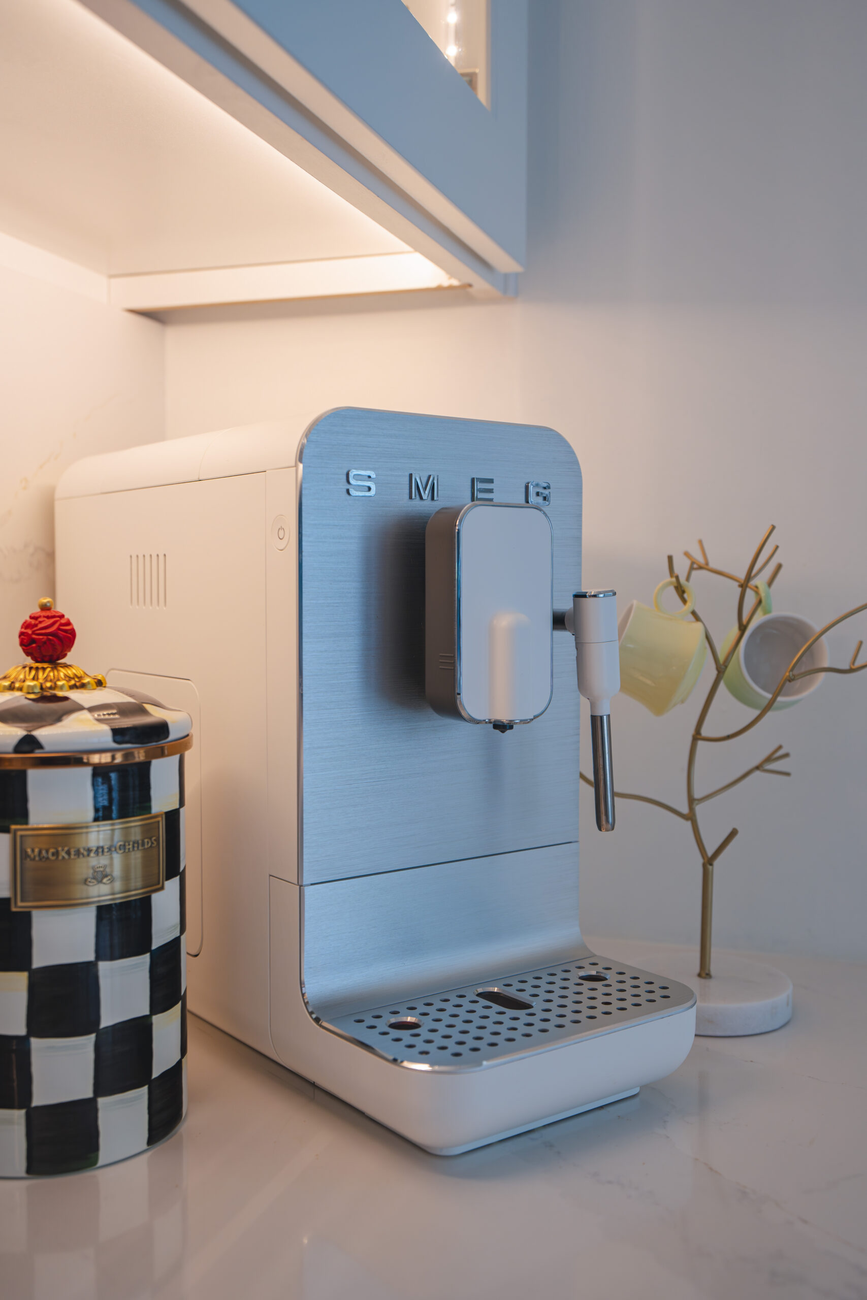A sleek white Smeg coffee maker on a white quartz countertop under integrated cabinet lighting.