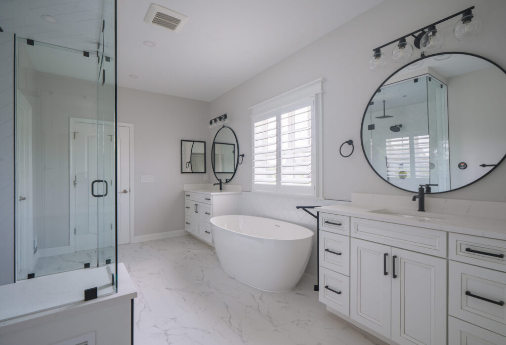 Large master bathroom remodel with a white shaker-style double vanity, round black-rimmed mirrors, marble floors, and a center soaking tub.