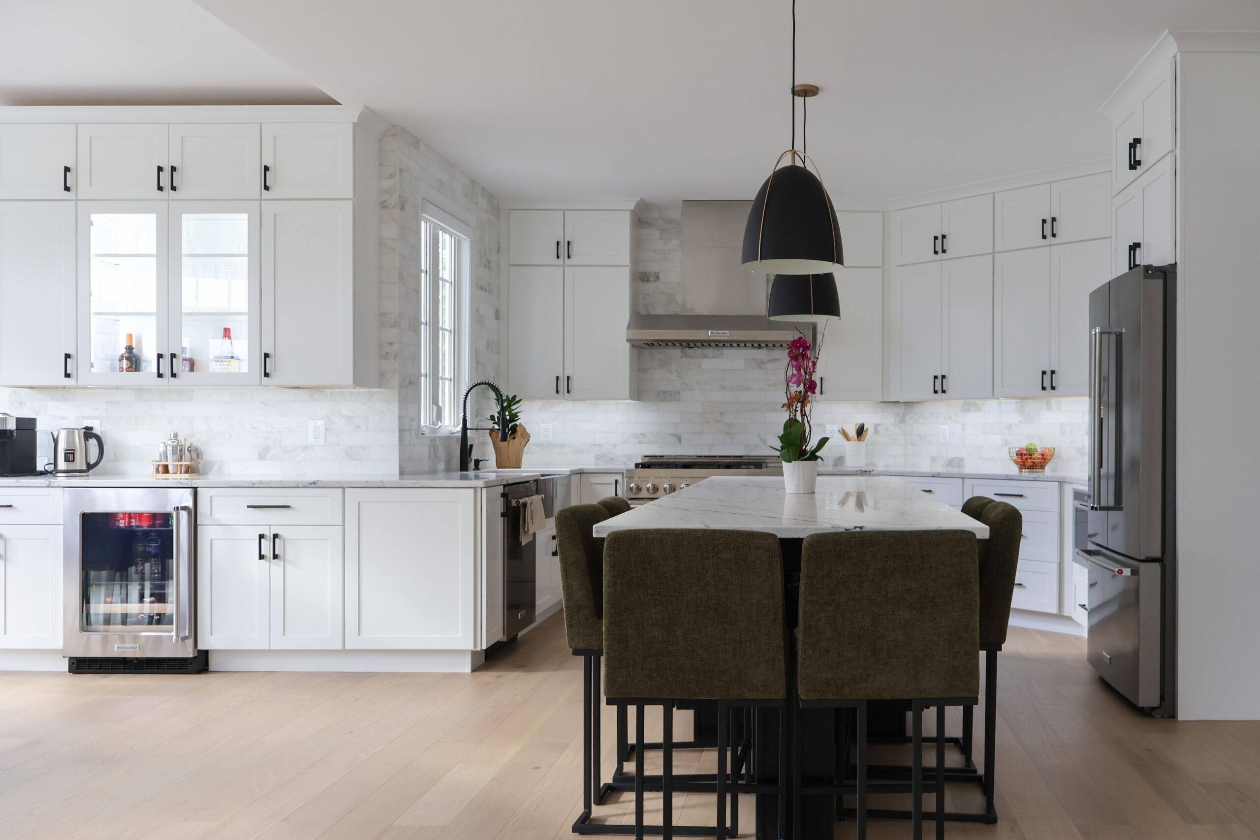 Open concept kitchen remodel in Washington DC with all-white shaker cabinets, matte black hardware, full-height marble tile backsplash, large marble island with green bar stools, wine fridge, and stainless steel appliances