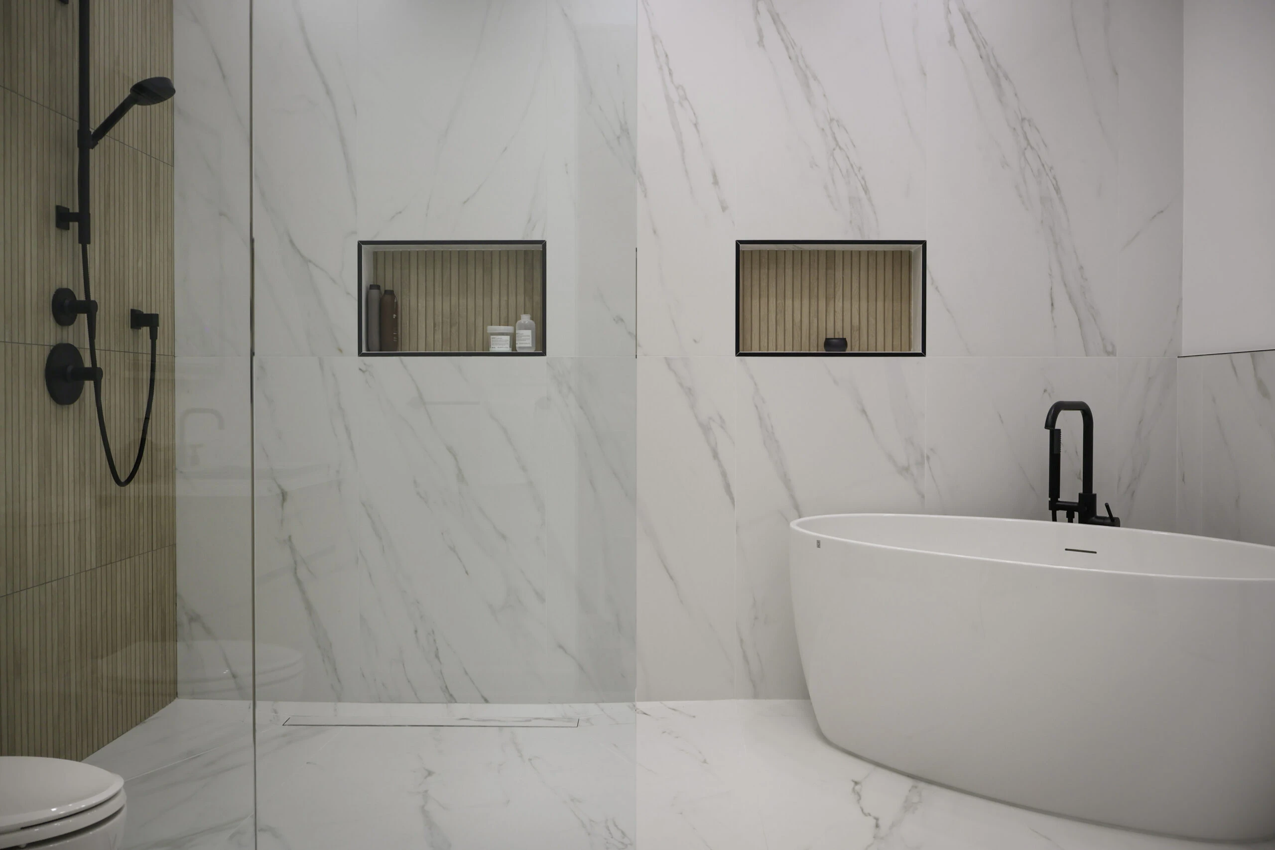 Spa bathroom remodel in McLean, VA with floor-to-ceiling marble slab wall cladding, curbless walk-in shower with linear drain and matte black fixtures, freestanding soaking tub with floor-mount faucet, and warm-toned niche inserts by Build Design Center