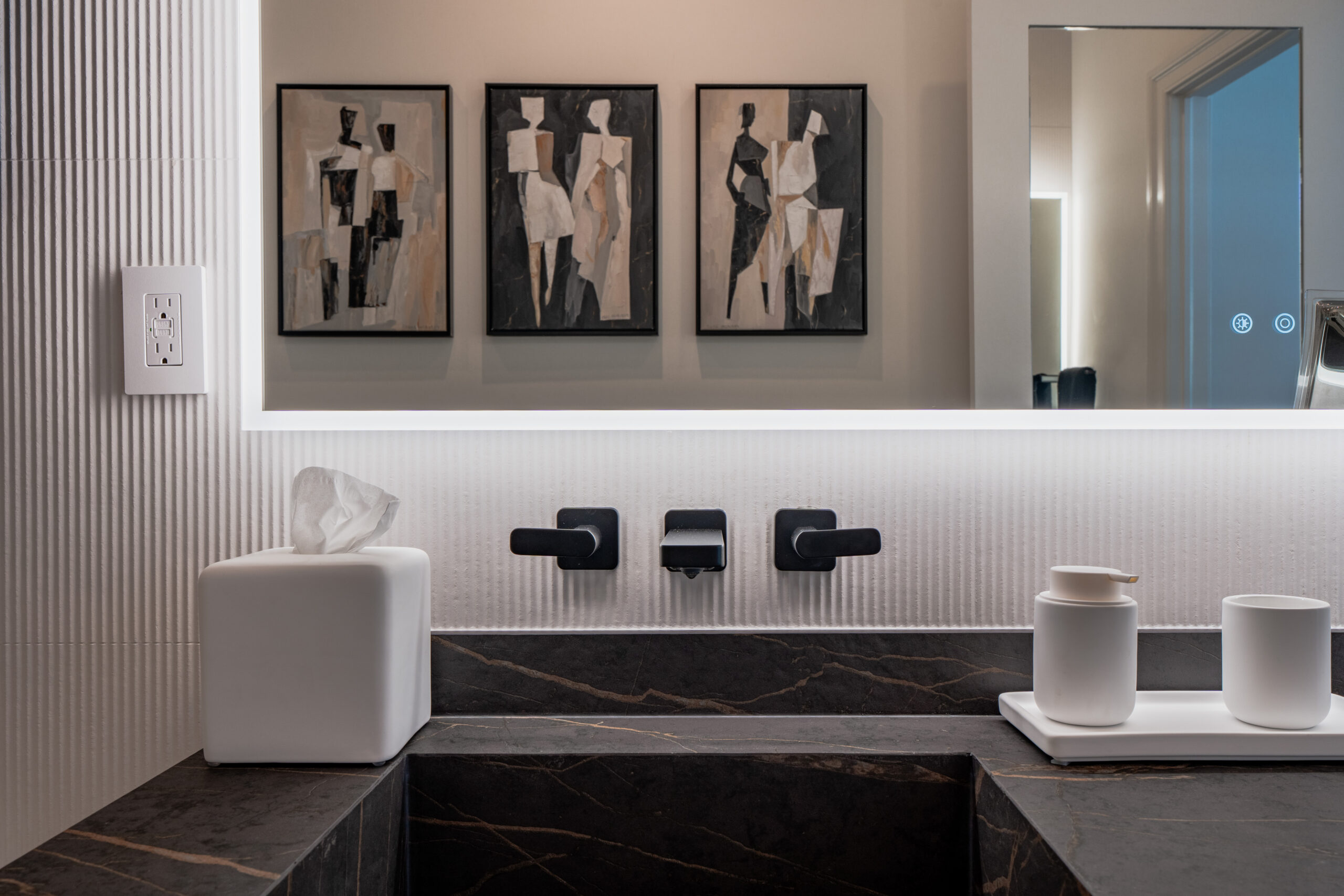 Black marble double vanity with matte black wall-mounted faucet, ribbed white tile backsplash, large backlit LED mirror reflecting triptych figurative artwork above, and white bathroom accessories in Washington DC luxury bathroom remodel