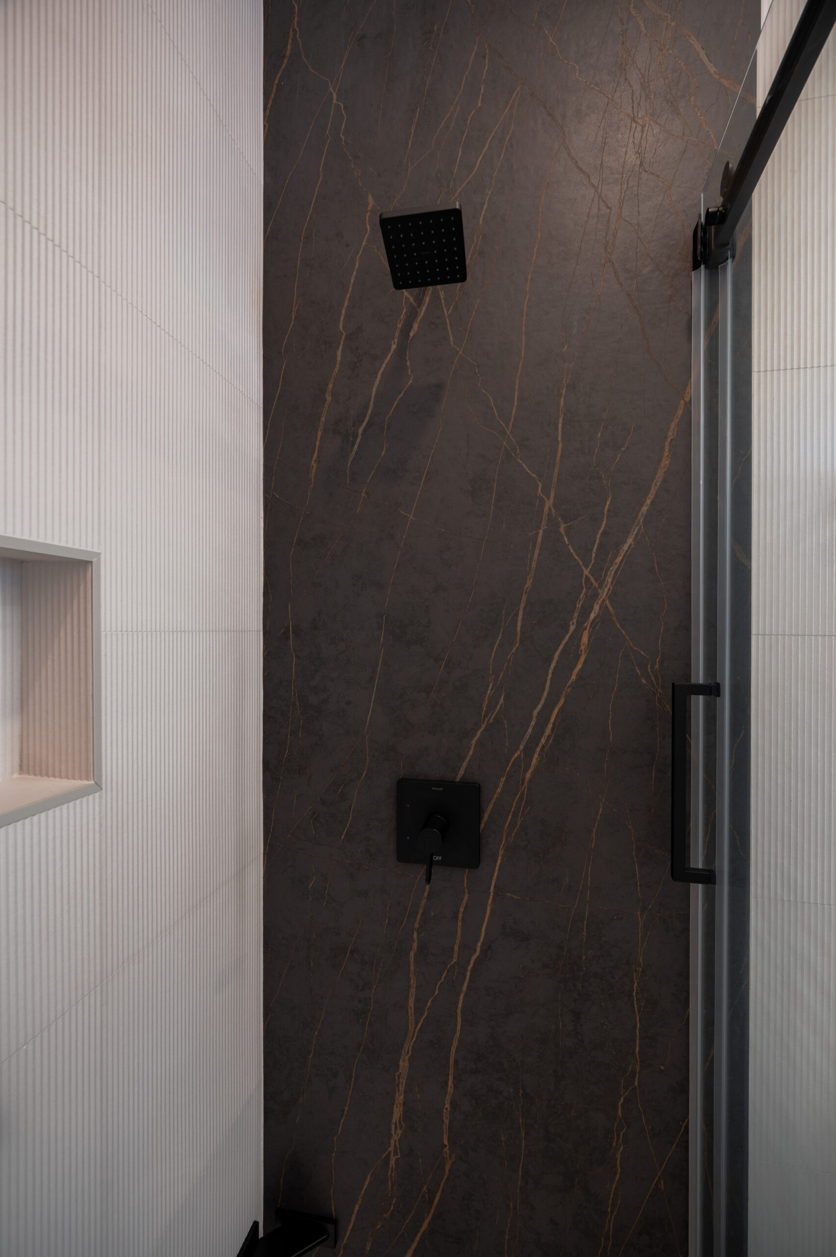 Close-up of dark marble with gold veining shower wall featuring matte black square rain shower head and thermostatic valve, with ribbed white tile accent wall and built-in shower niche in Washington DC bathroom remodel