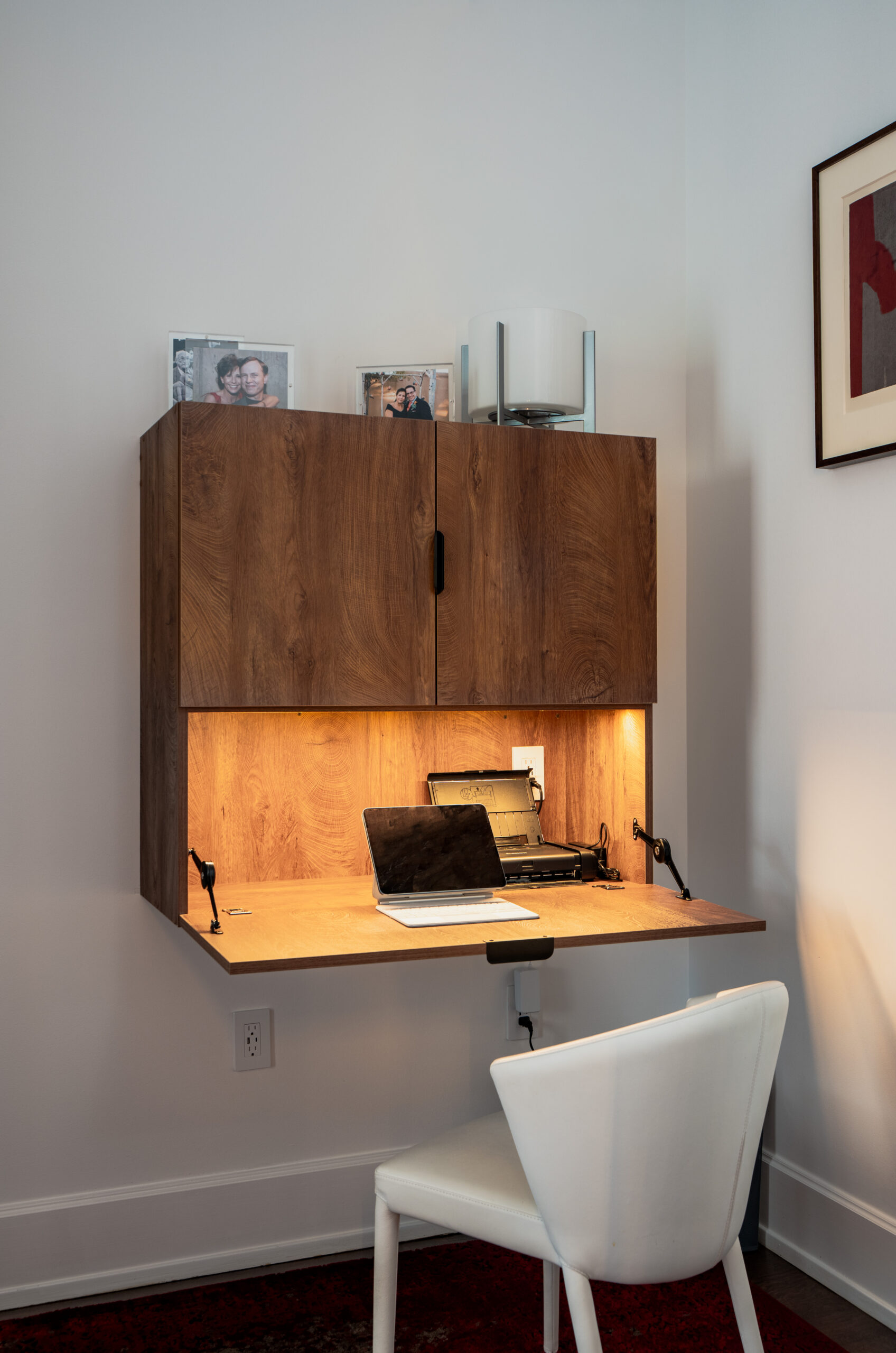 Close-up of built-in walnut fold-down desk with warm LED under-cabinet lighting, laptop, dual swing-arm task lamps, and upper storage cabinet in Washington DC custom home renovation