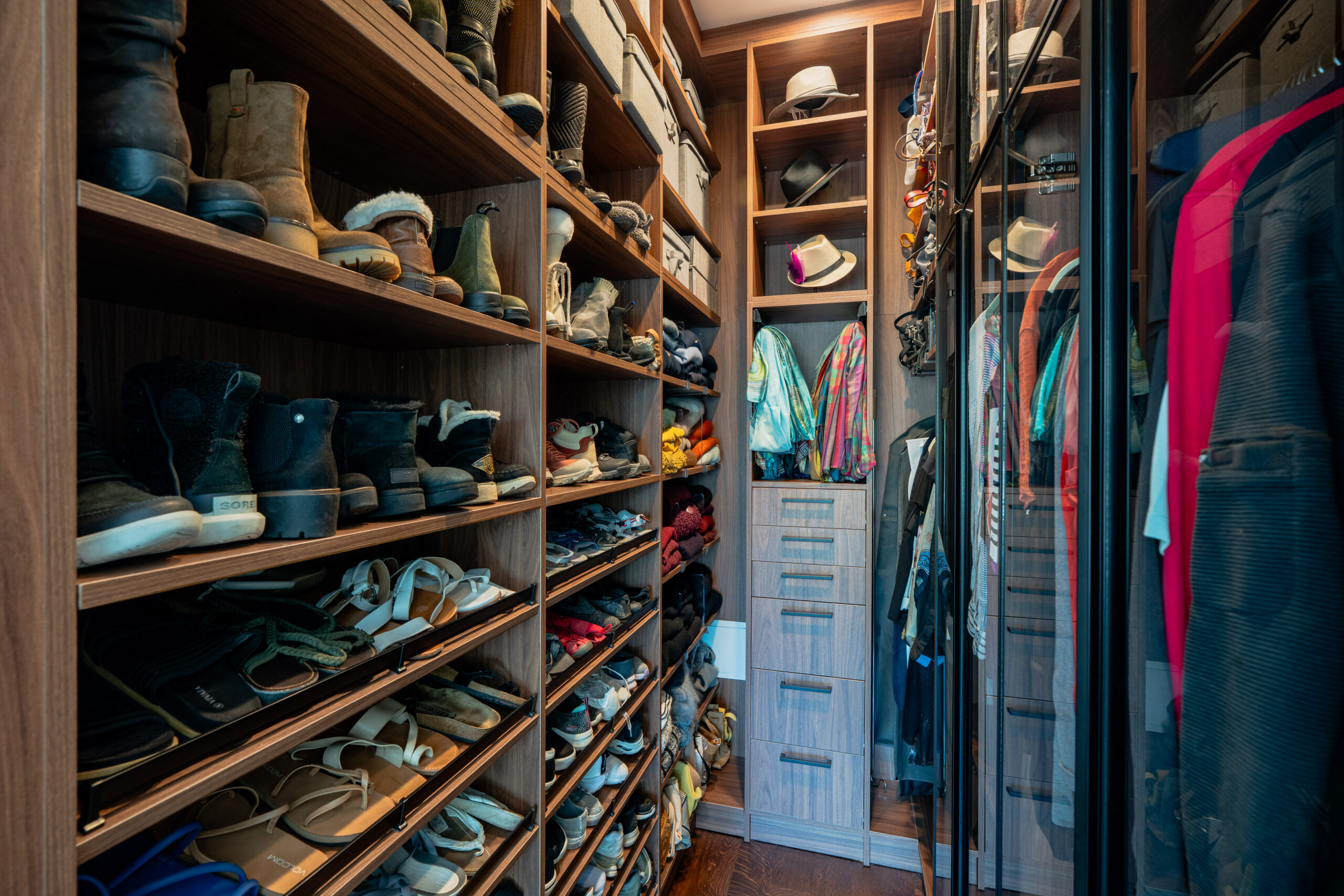 Custom walk-in closet showing angled walnut shoe wall shelving with boots and sandals, open hat display cubbies, built-in gray drawer tower, colorful hanging scarves, and black steel glass wardrobe doors in Washington DC luxury home renovation