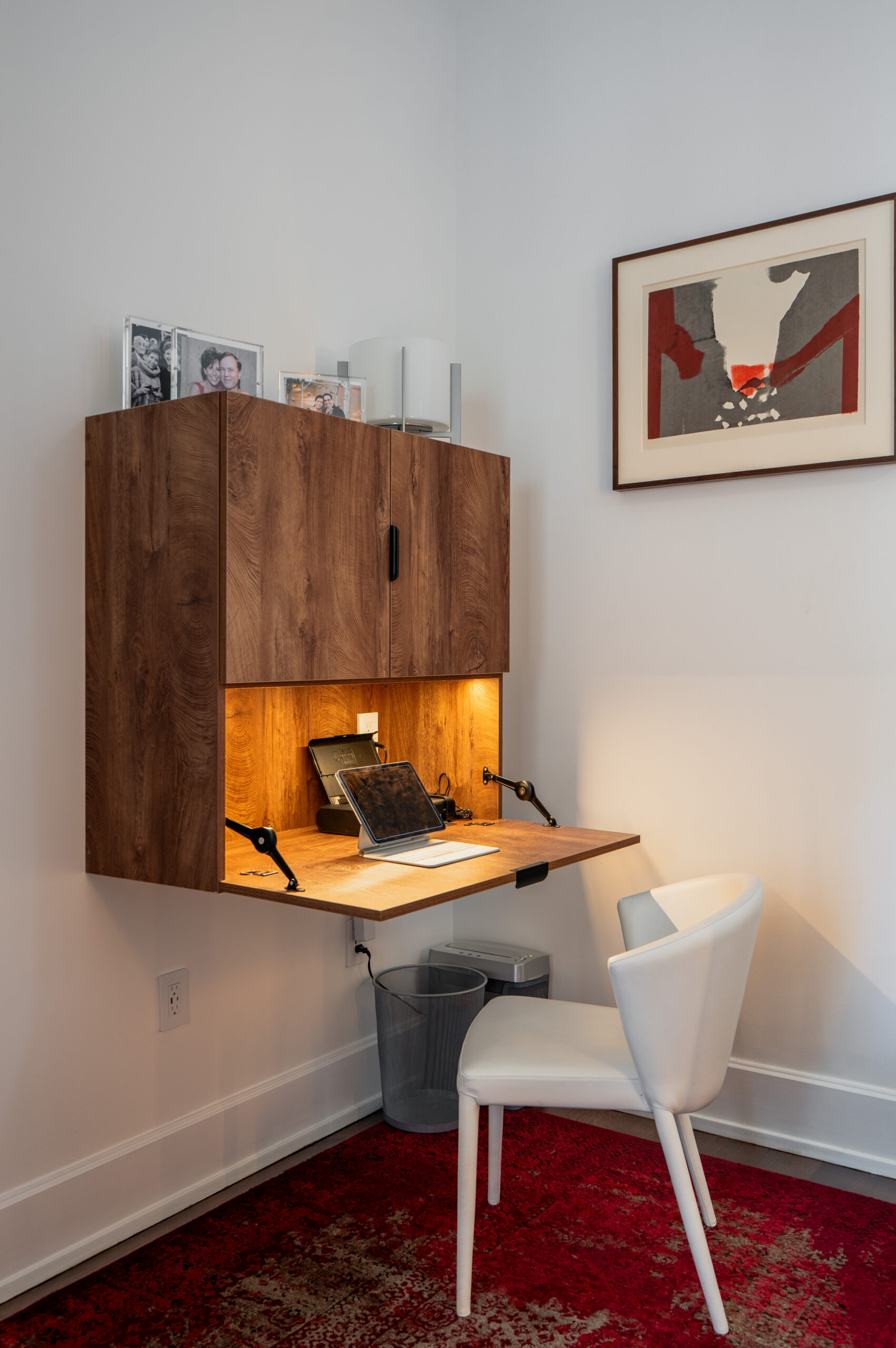 Custom walnut wall-mounted fold-down desk with built-in LED lighting, upper storage cabinet, white leather chair, and red abstract artwork in Washington DC home office