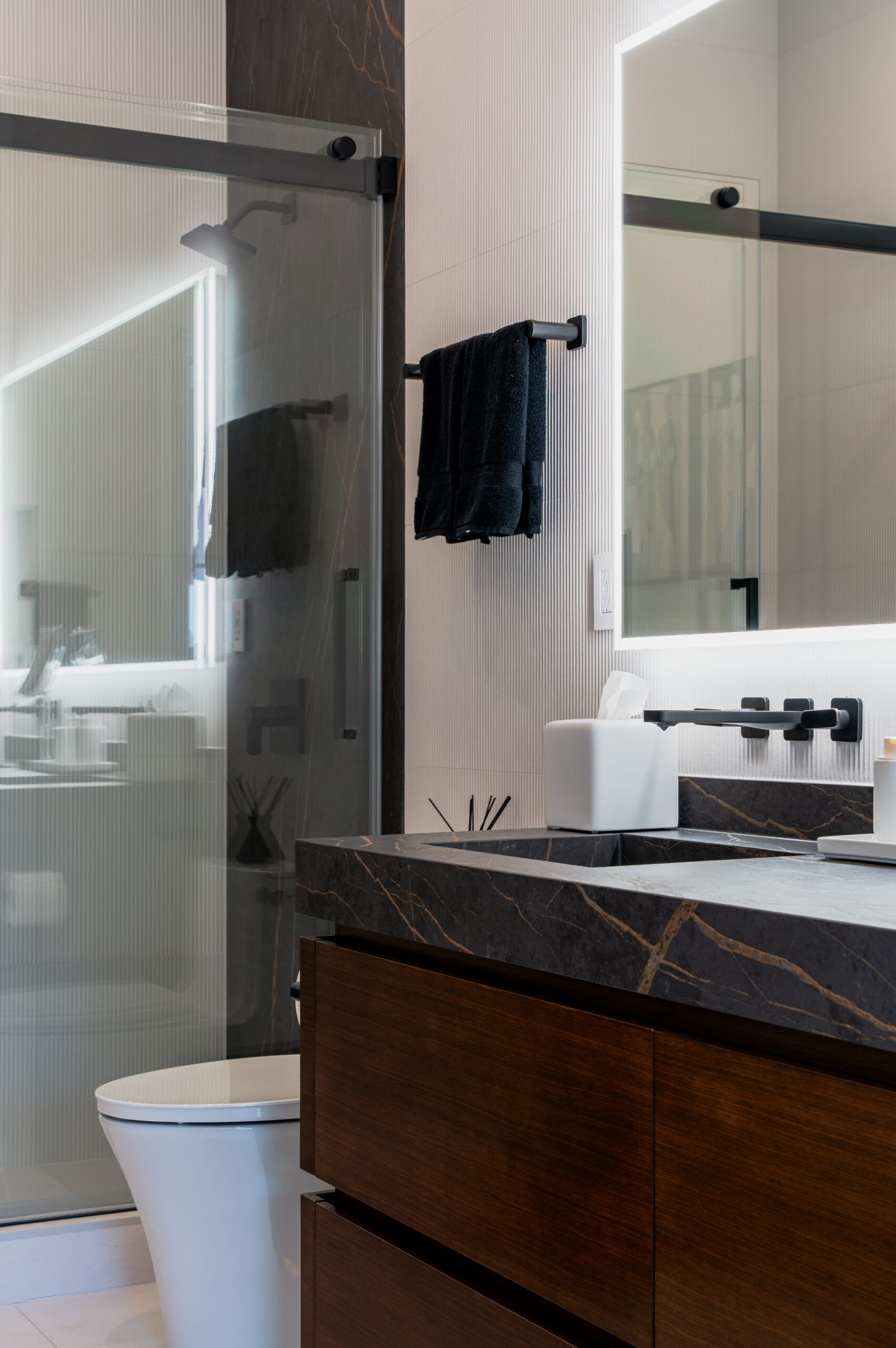 Floating walnut bathroom vanity with black marble countertop and undermount sink, matte black fixtures, backlit LED mirror, wall-mounted toilet, and frameless sliding glass shower enclosure with dark marble accent wall in Washington DC bathroom remodel